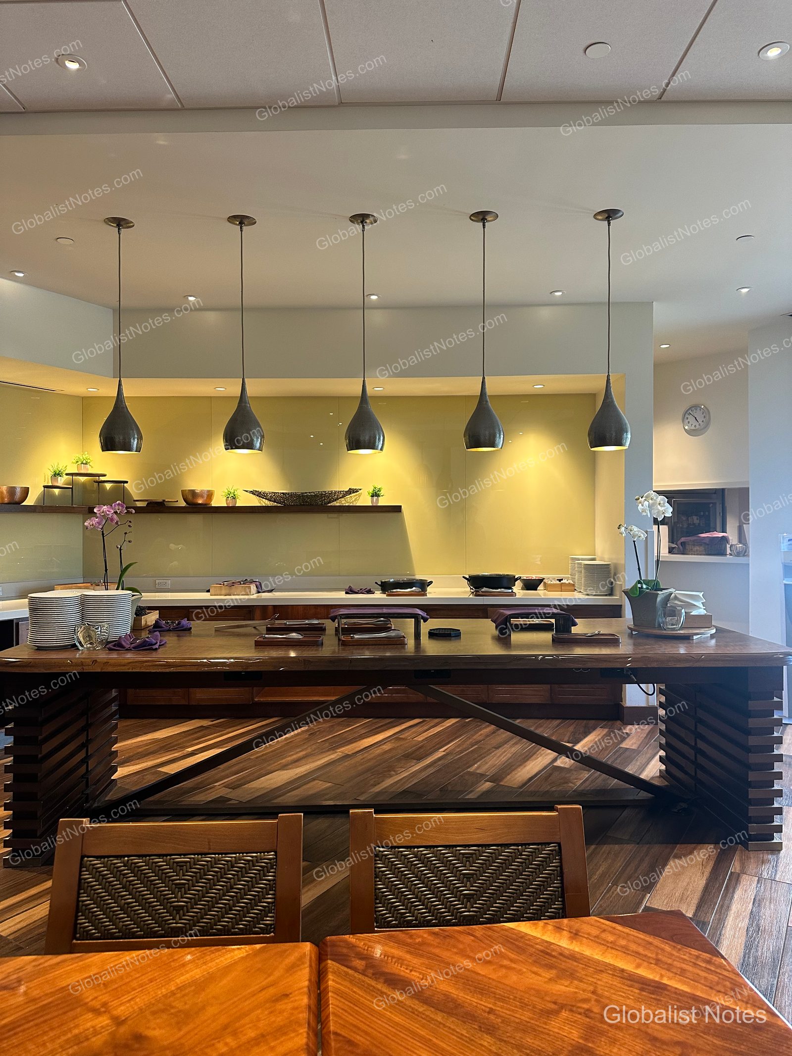 Interior view of the Hyatt Regency Maui Resort and Spa in Lahaina, Hawaii, featuring five pendant lights hanging low over a long wooden table. In the background, illuminated wall shelves and neatly arranged tableware create a calm, organized atmosphere.