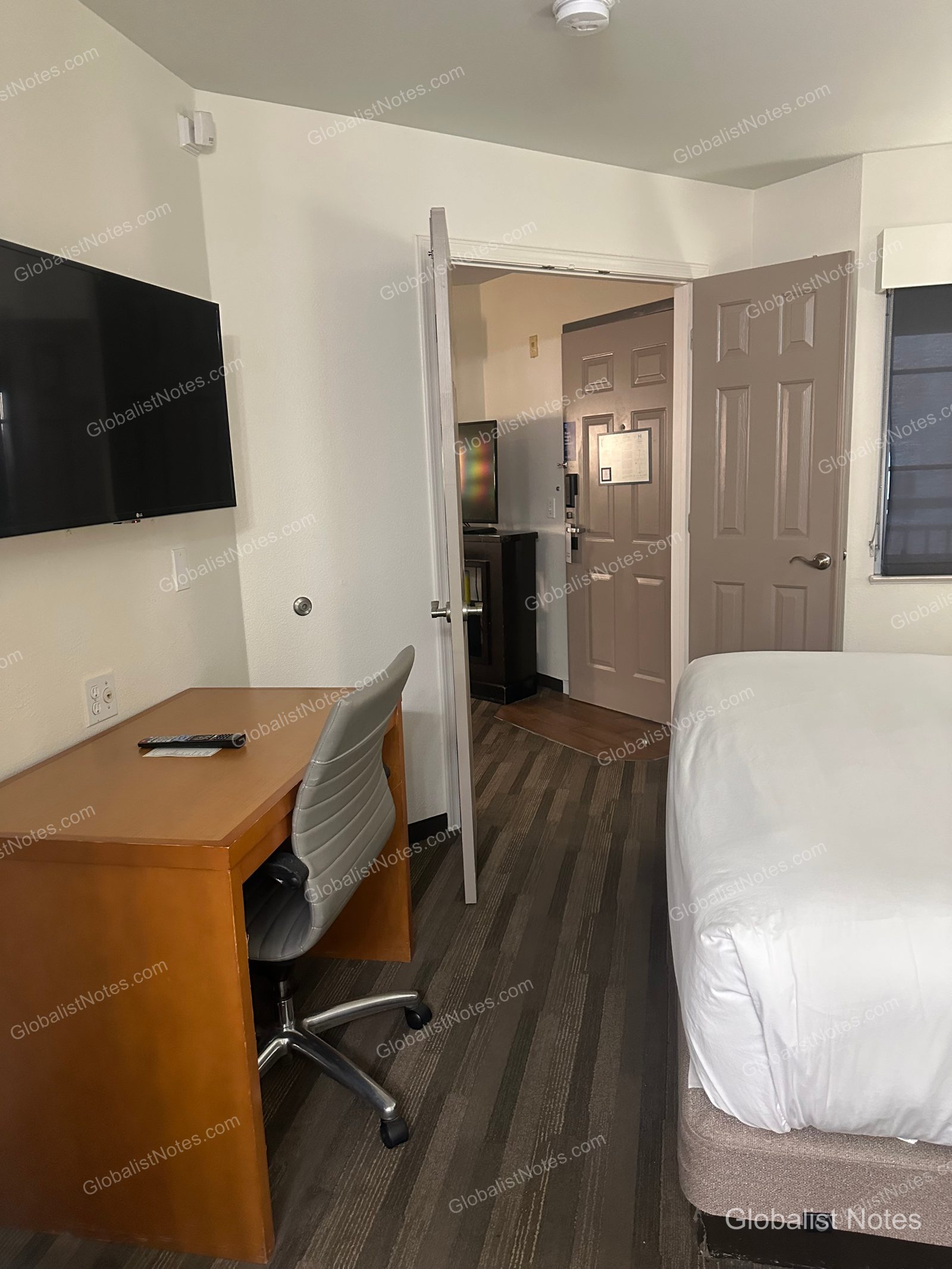 Hyatt House Scottsdale/Old Townのワンベッドルームスイートの室内。左手に壁掛けテレビとデスク、右手にベッドの一部が見え、開いたドアの向こうに玄関と廊下が続いている。