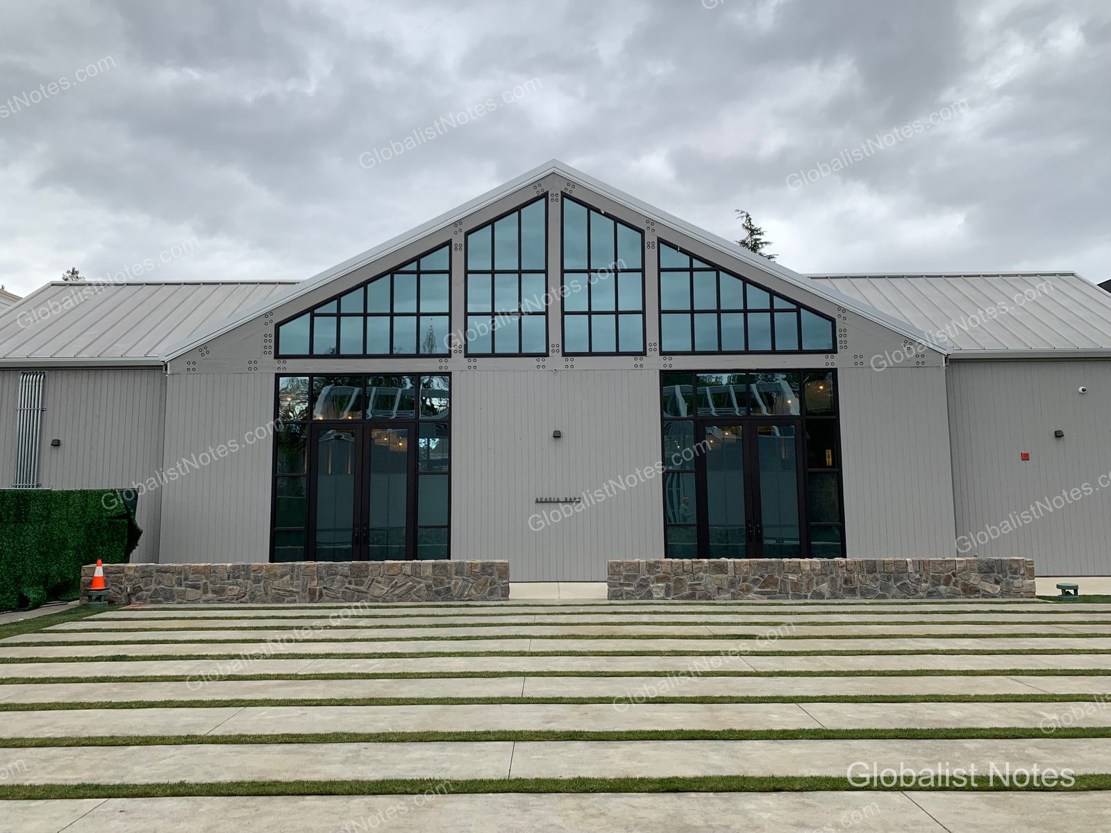 Exterior of a modern gabled building at Alila Napa Valley in St. Helena, USA. In front of the gray walls with large glass windows, there is an outdoor space with stone-walled planters and a concrete patio with grass patches under a cloudy sky.