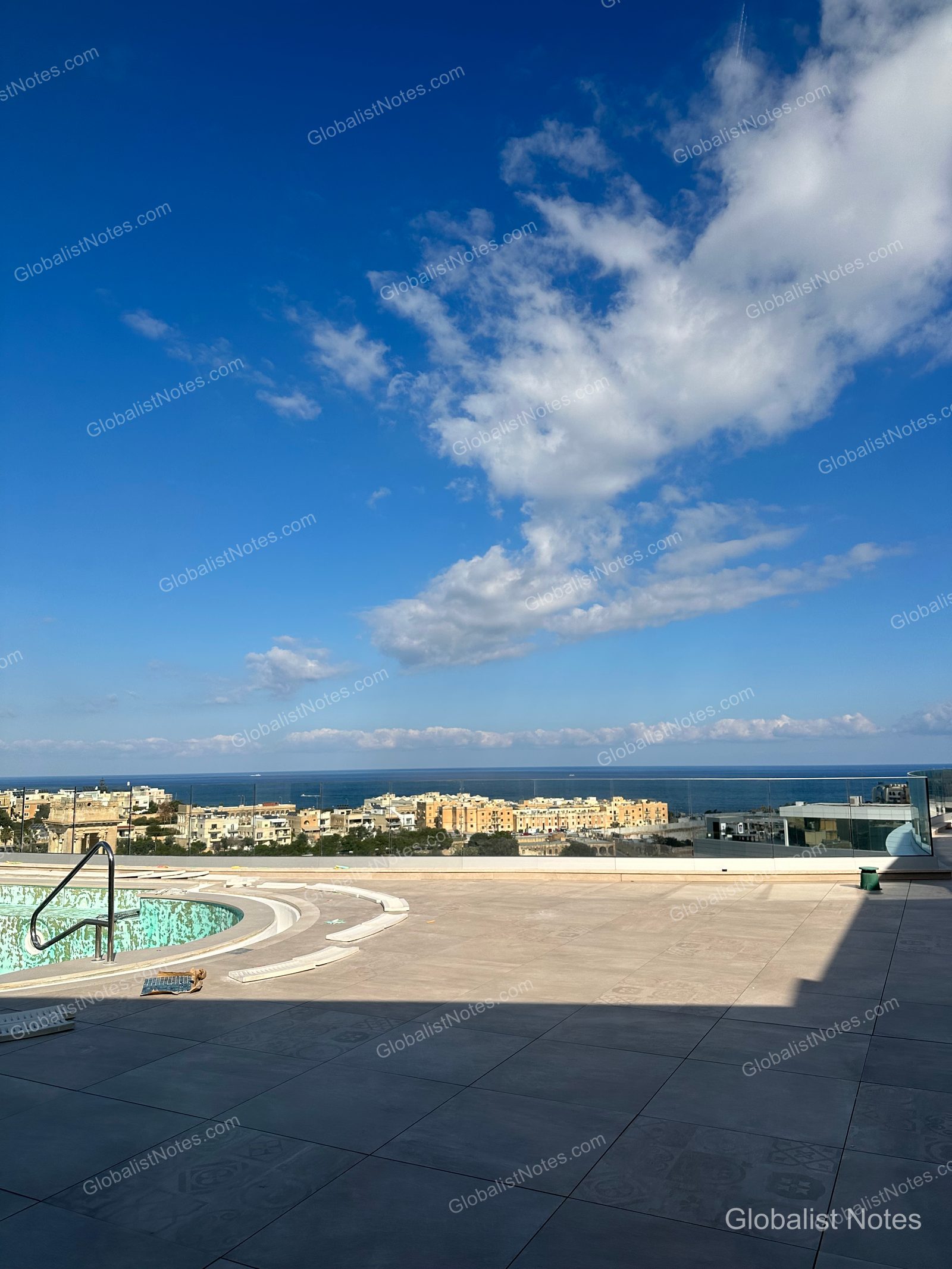 View from the outdoor pool deck at Hyatt Regency Malta, looking past glass railings toward the St. Julian's cityscape and the Mediterranean Sea.