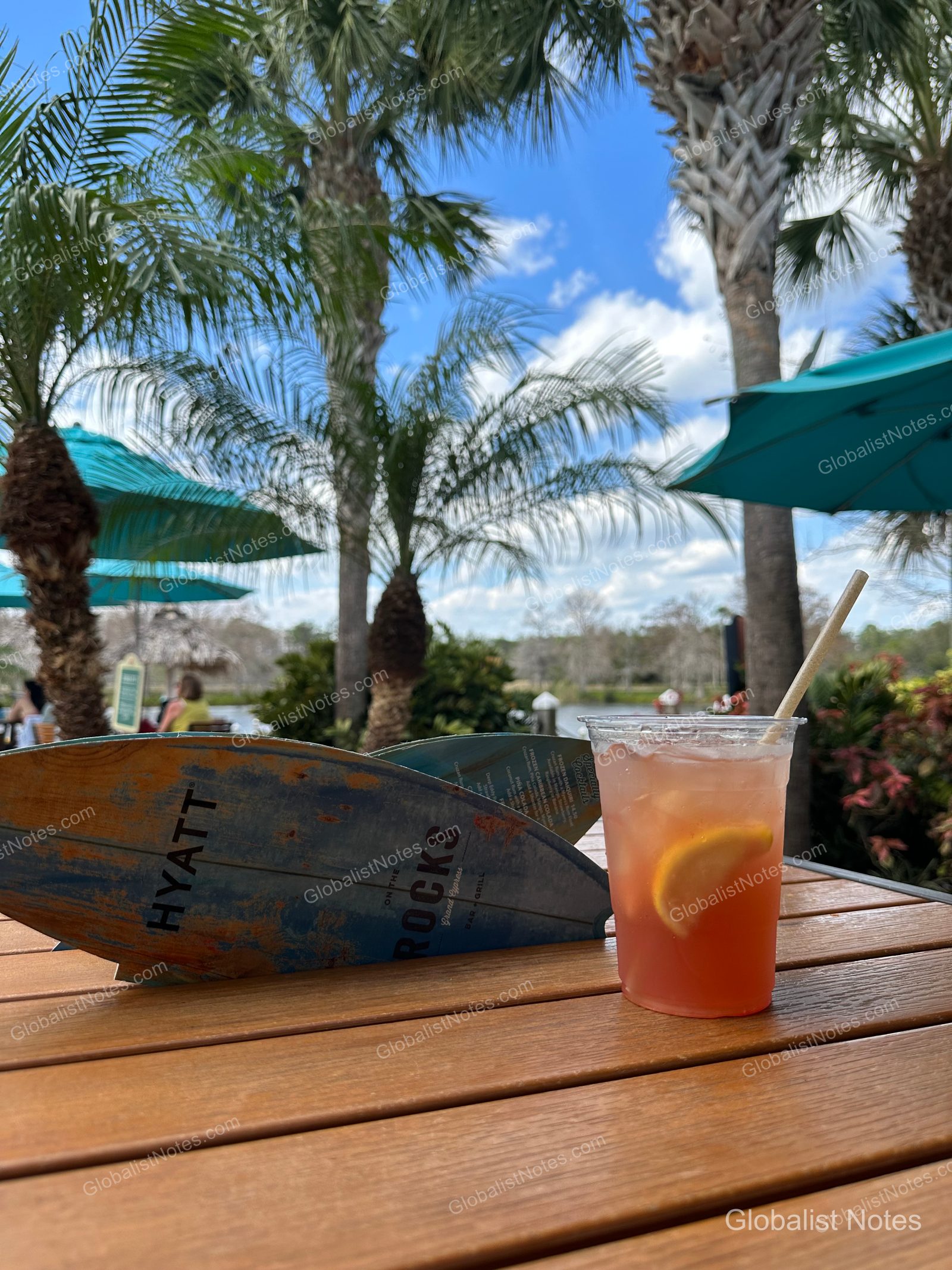 A cocktail and the 'On the Rocks' bar menu on a table by the outdoor pool at Hyatt Regency Grand Cypress, with palm trees, blue sky, and blue umbrellas in the background.