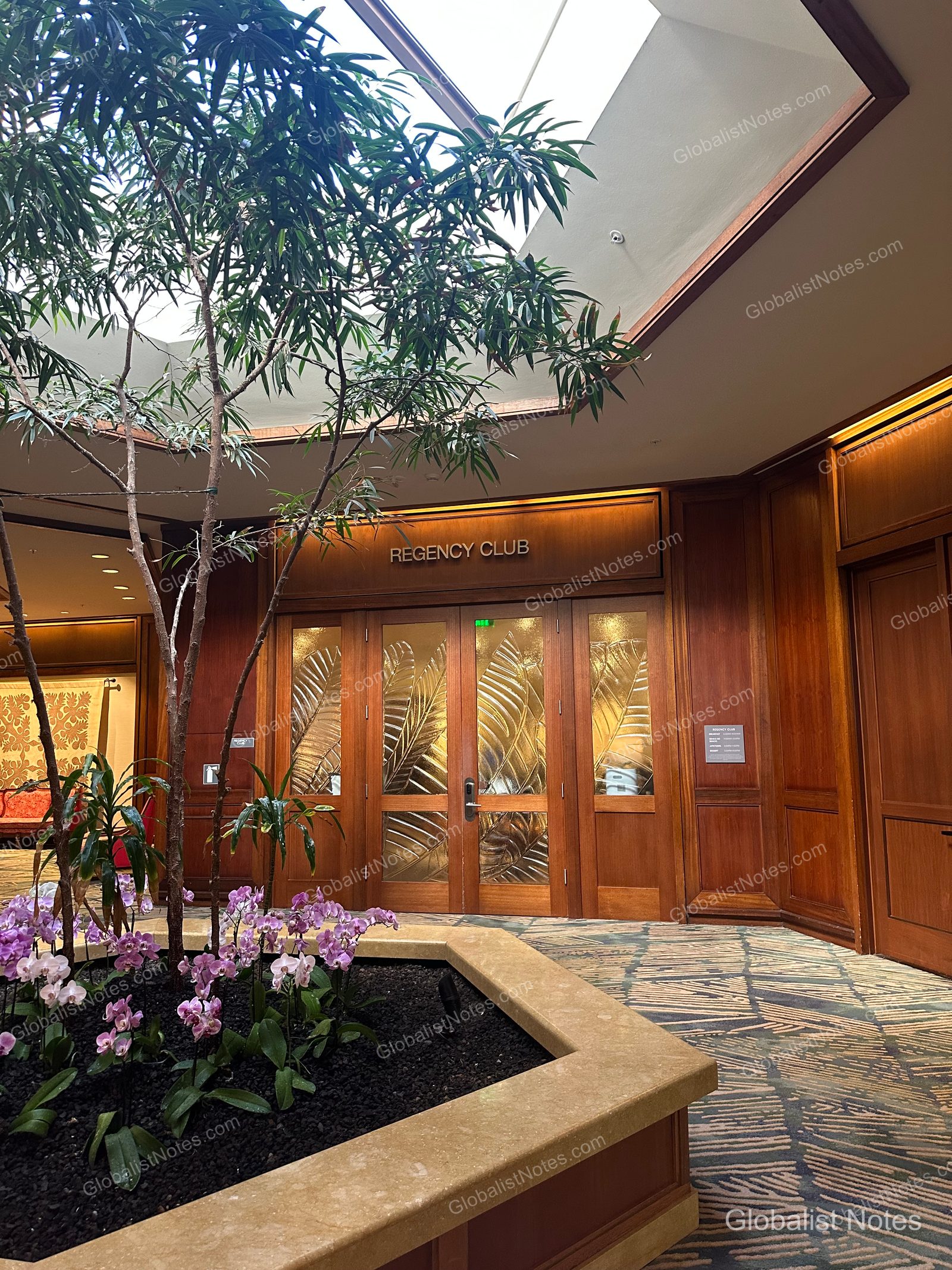 Entrance to the Regency Club at Hyatt Regency Maui Resort and Spa. A planter with trees and purple flowers sits beneath a ceiling skylight, leading to the glass doors of the Regency Club featuring a leaf-patterned design.