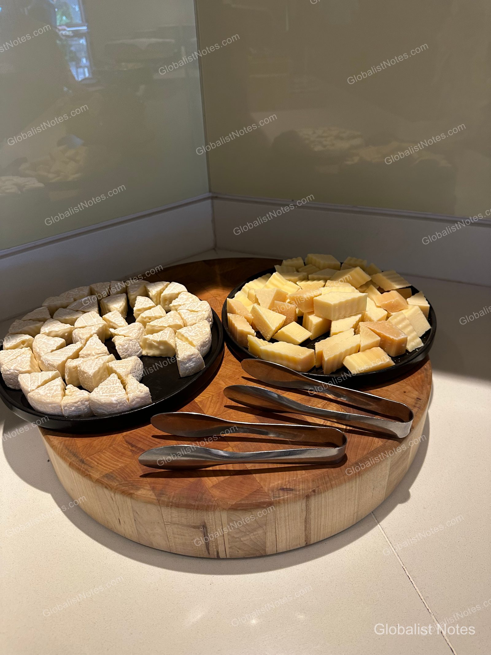 Two black plates on a round wooden board in the Hyatt Regency Maui Resort and Spa lounge, each holding bite-sized portions of soft and hard cheeses, with serving tongs placed alongside.