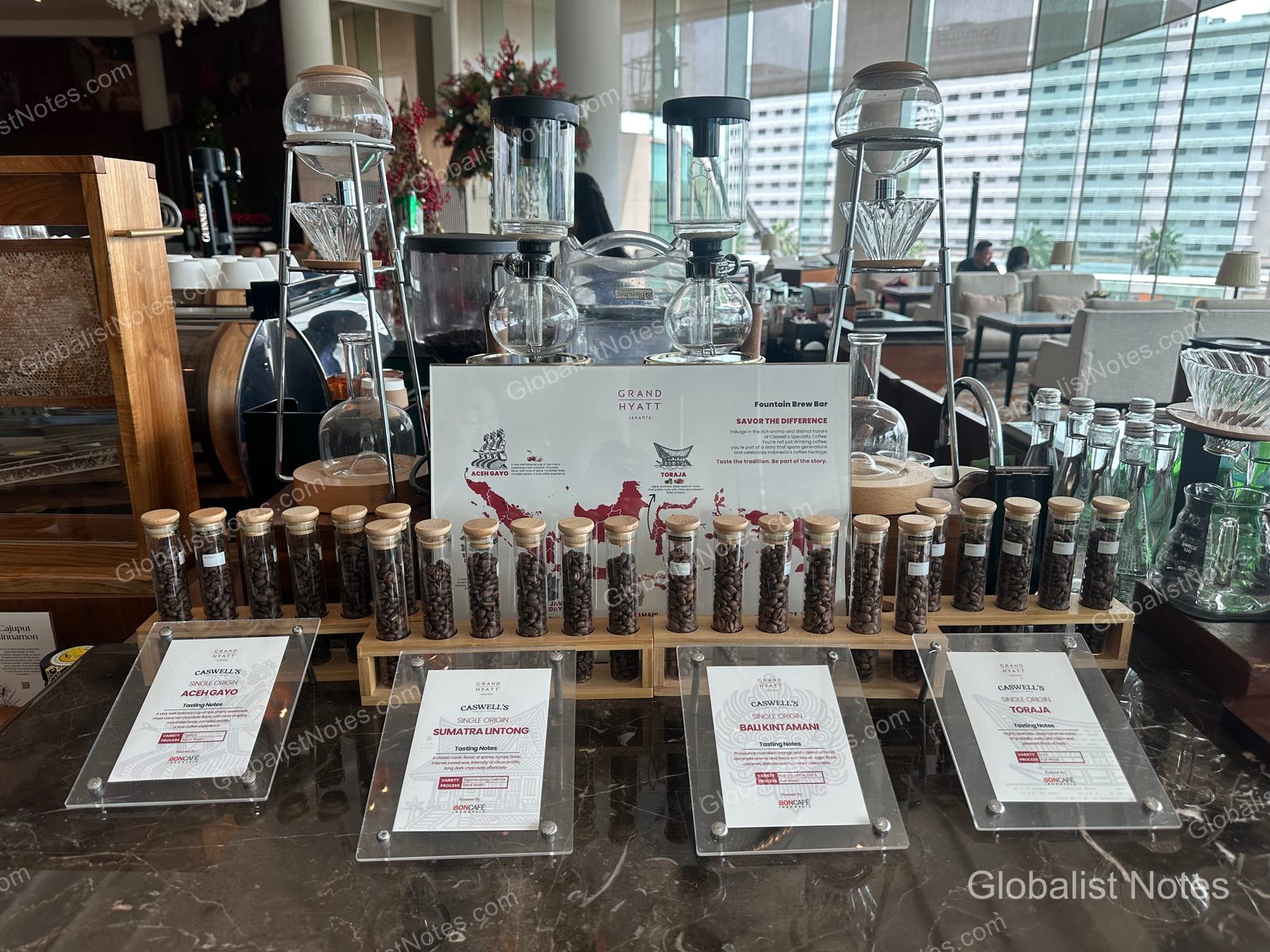 A display of various Indonesian coffee bean samples at the Fountain Brew bar in Grand Hyatt Jakarta. Glass tubes hold the beans, accompanied by information cards for regional varieties like Aceh Gayo, Sumatra Lintong, Bali Kintamani, and Toraja, along with a siphon brewing setup.