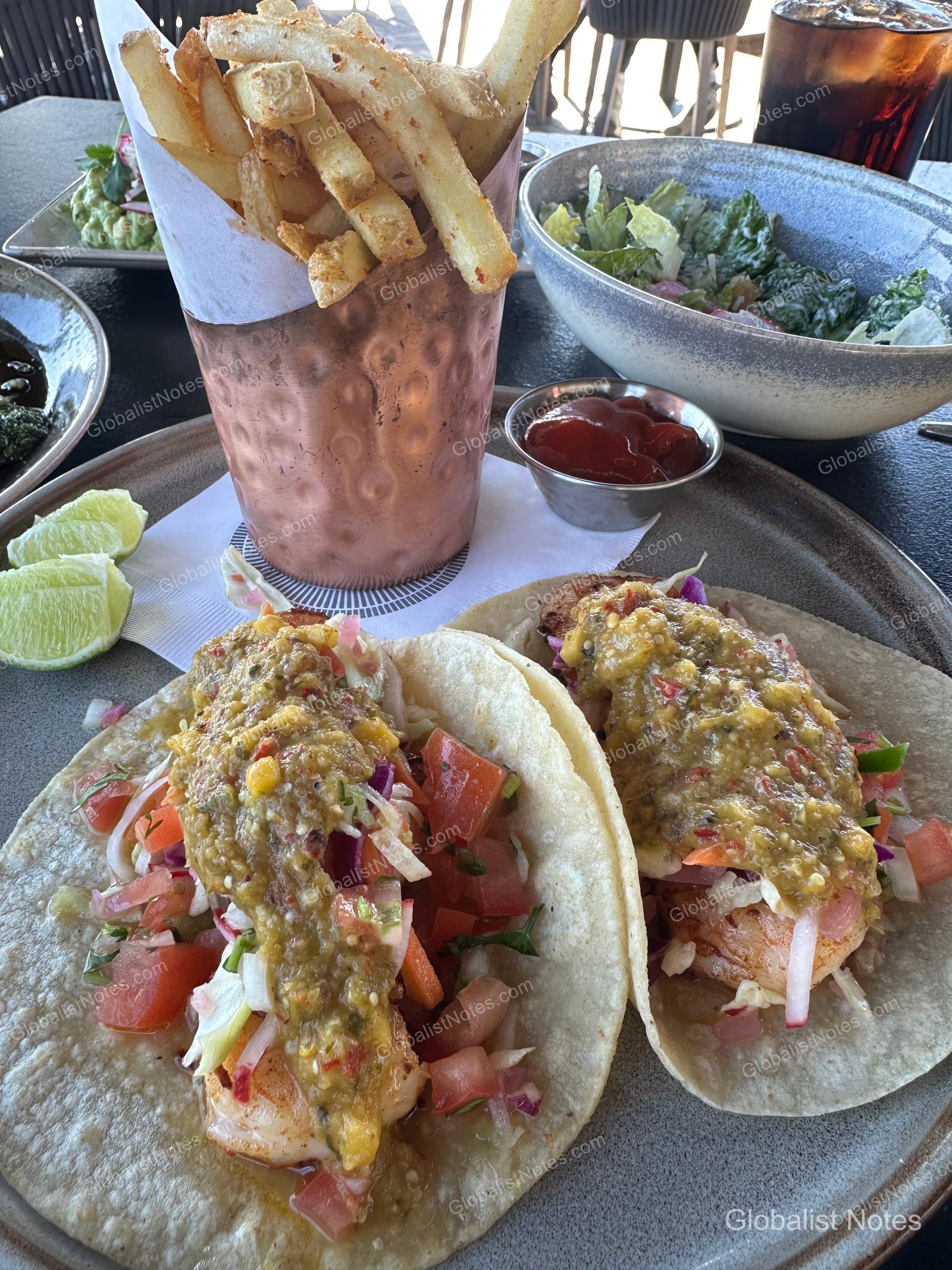 A lunch served at Alila Ventana Big Sur featuring two tacos topped with grilled shrimp and fresh salsa, served with a side of fries in a copper cup, a salad, and ketchup on a table.