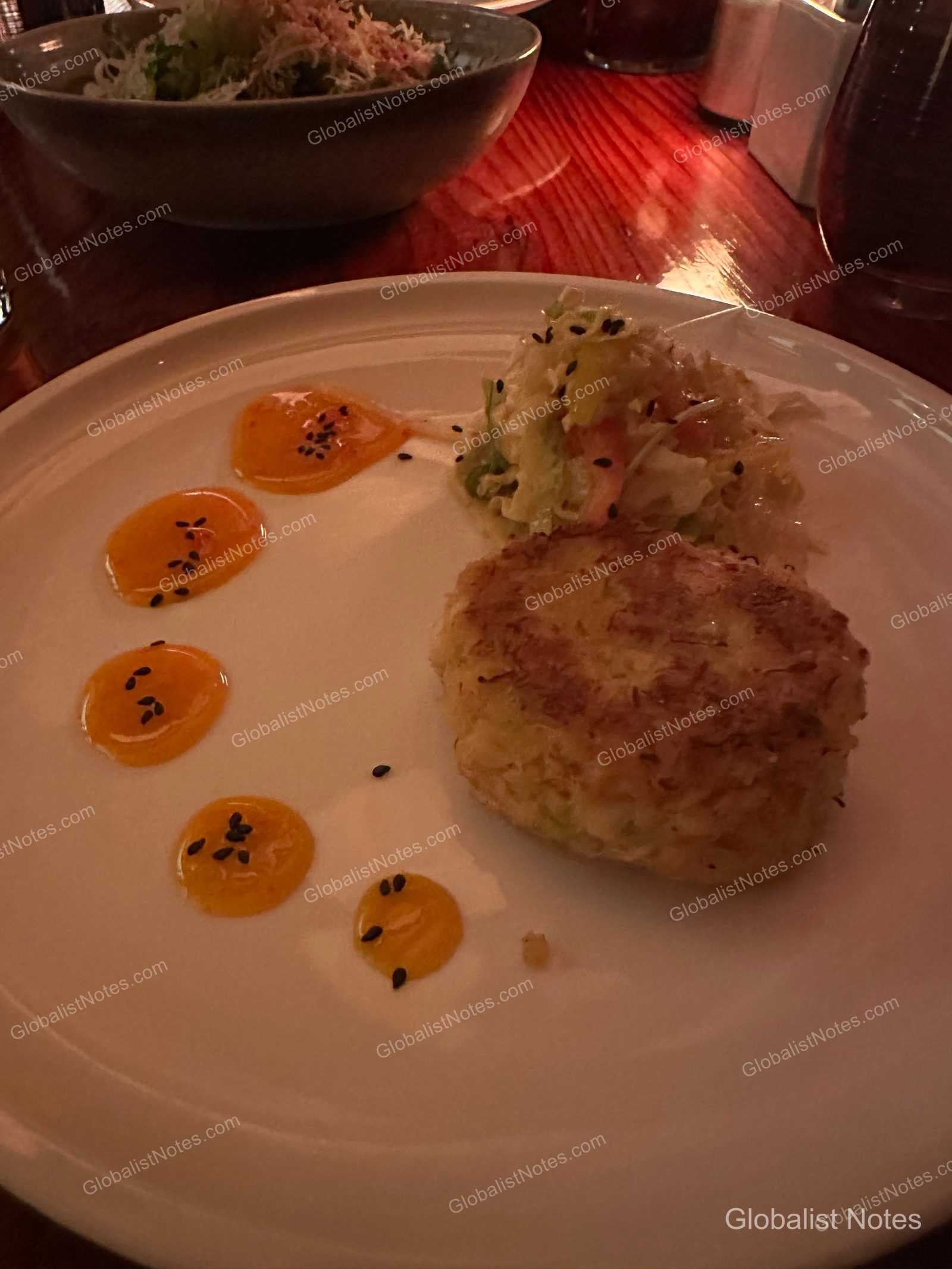 A dinner dish at Alila Ventana Big Sur featuring a crab cake and salad on a white plate, garnished with five dots of yellow sauce. A wooden table and other tableware are visible in the blurred background.