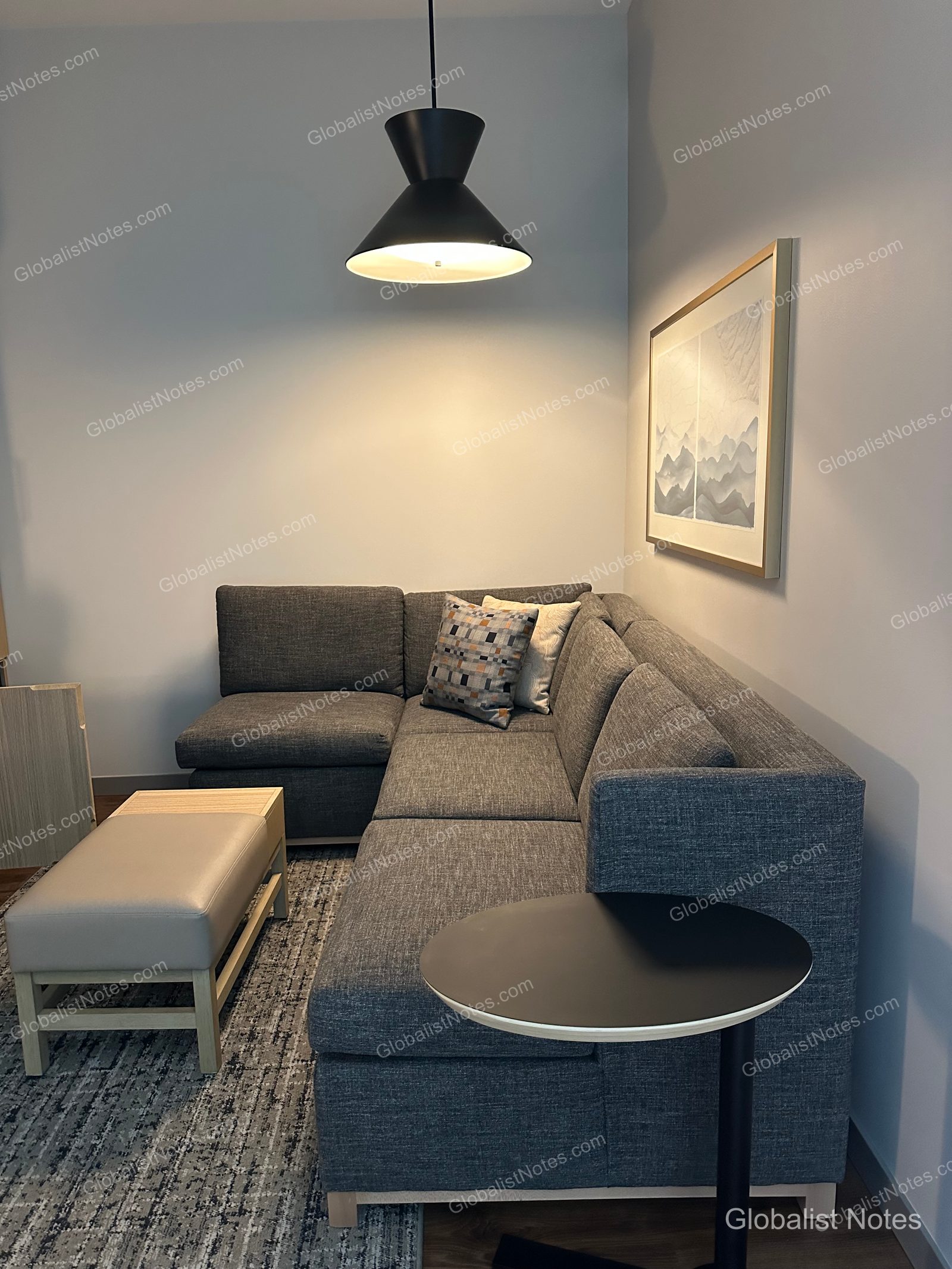 Hyatt Place Bozeman Yellowstone Airportのジュニアスイートルームのリビングエリア。グレーのL字型ソファ、ベージュのオットマン、黒い円形のサイドテーブルが置かれ、壁には山の絵が飾られています。