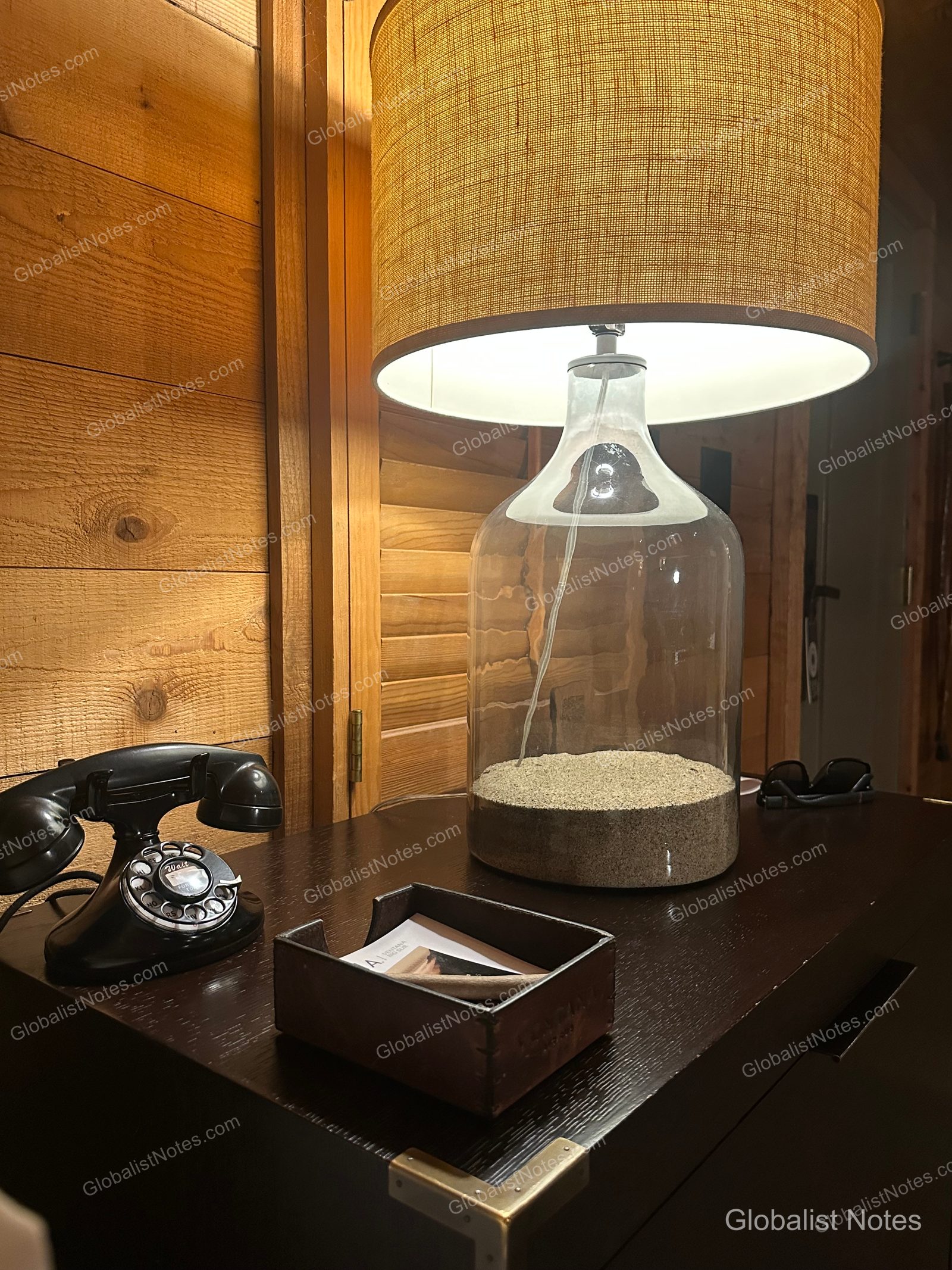 A vintage-style black rotary phone, a sand-filled glass lamp, and a leather notepad holder placed on dark wooden furniture in an Alila Ventana Big Sur Junior Suite.