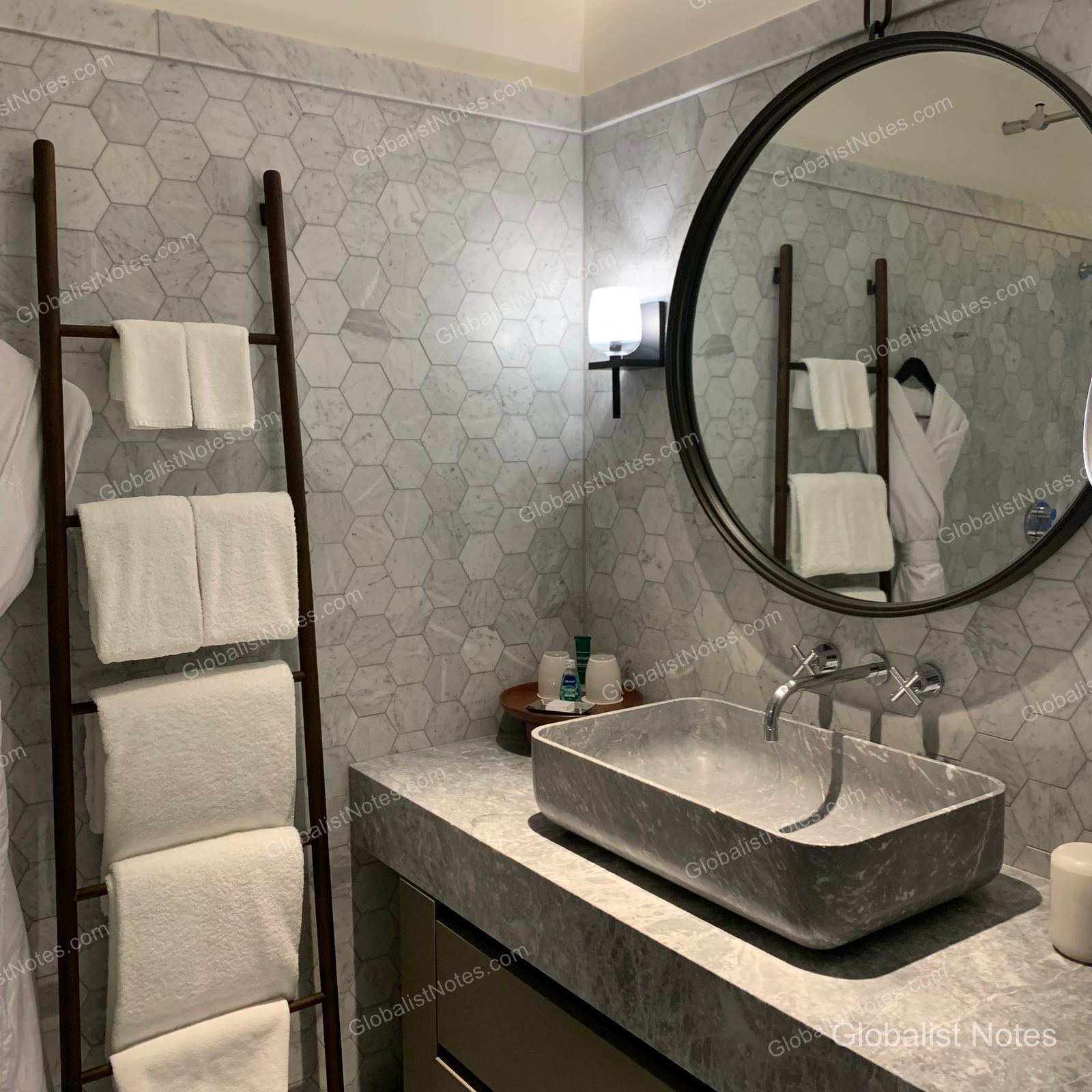 Bathroom interior at Alila Napa Valley, featuring hexagonal marble tile walls, a circular mirror, and a square vanity sink. A ladder-style towel rack stands on the left, and a bathrobe is reflected in the mirror.