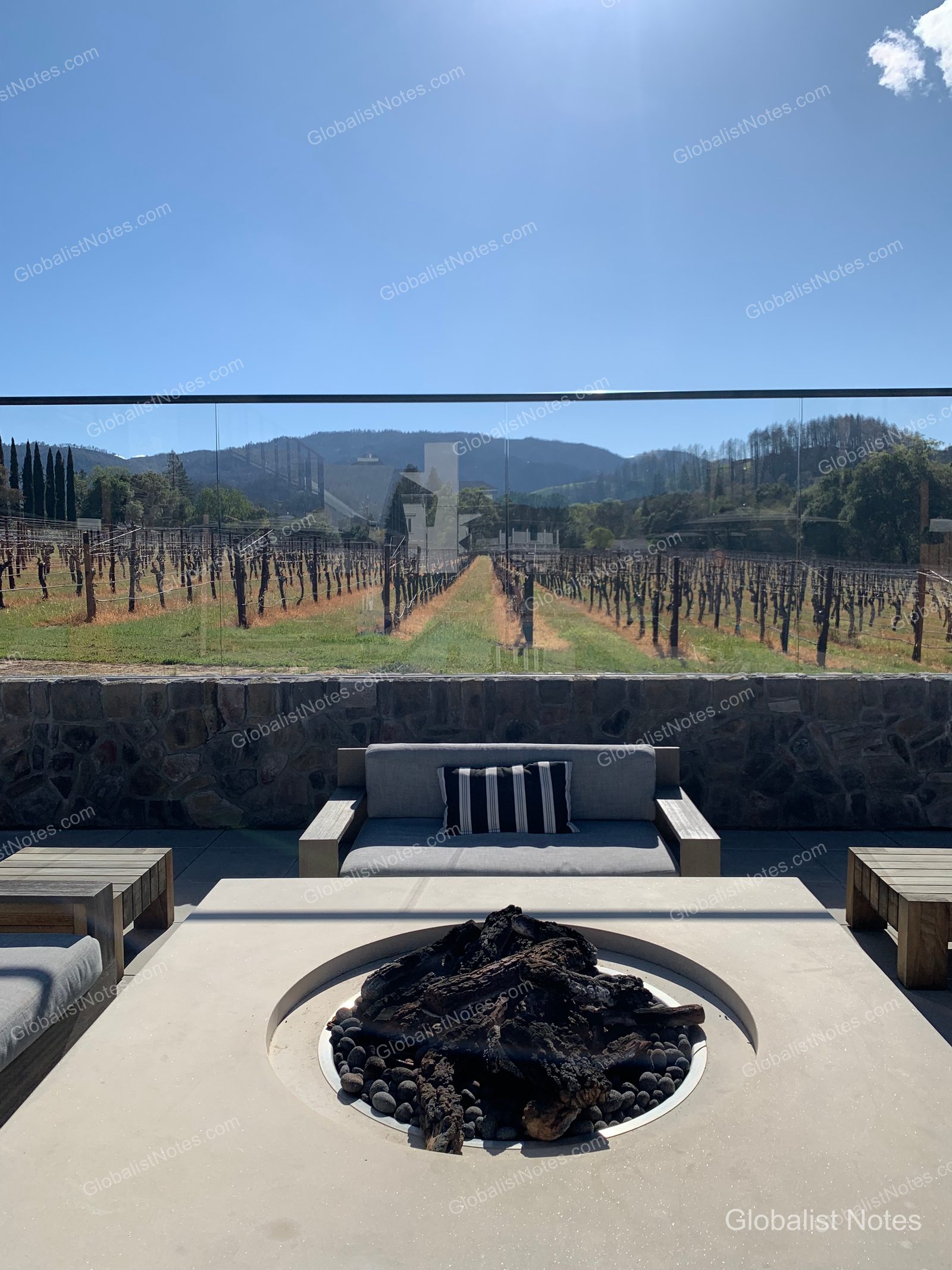 View from the balcony of a guest room at Alila Napa Valley in St. Helena, California. The scene shows a terrace equipped with a fire pit and sofa, overlooking the Napa Valley vineyards and distant mountain ranges on a clear day.