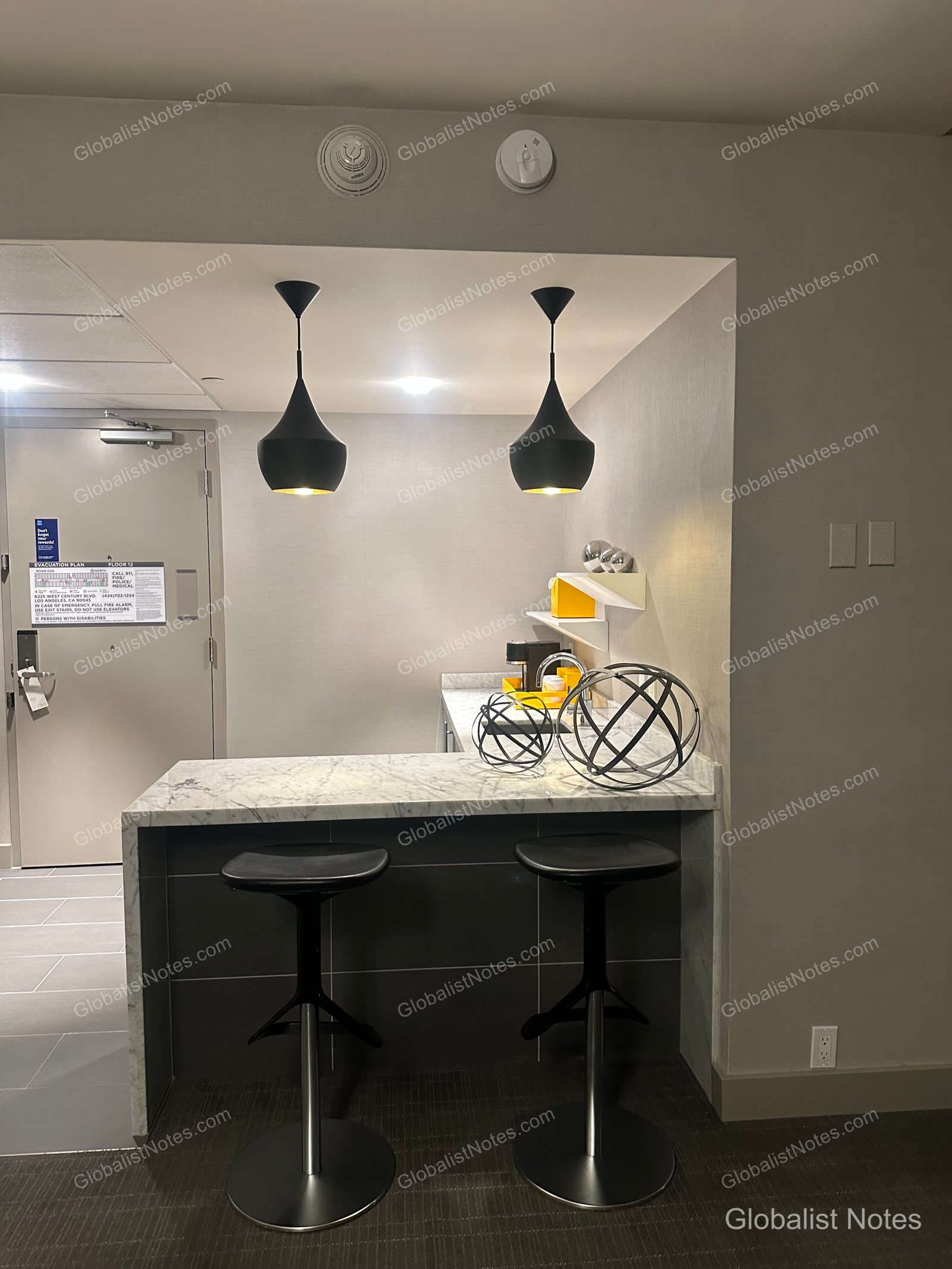 The bar area inside a One Bedroom King Suite at Hyatt Regency Los Angeles International Airport. It features a marble-topped table with two black bar stools and two modern black pendant lights overhead. A spherical decorative object and a coffee machine are placed on the table.