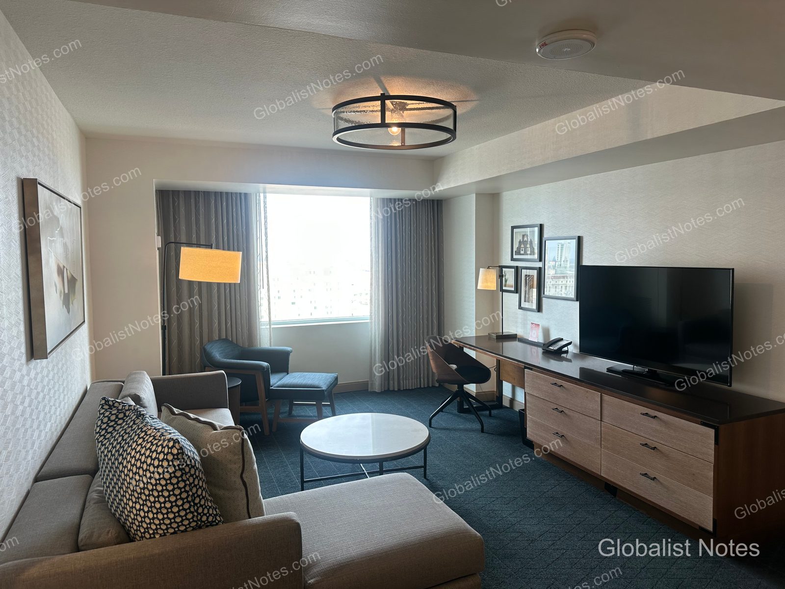 Living area of an Executive Suite at the Grand Hyatt San Antonio River Walk. It includes a gray sofa, a round table, an armchair with an ottoman by the window, a wall-mounted TV, and a long desk, all set in a modern, neutral-toned interior.