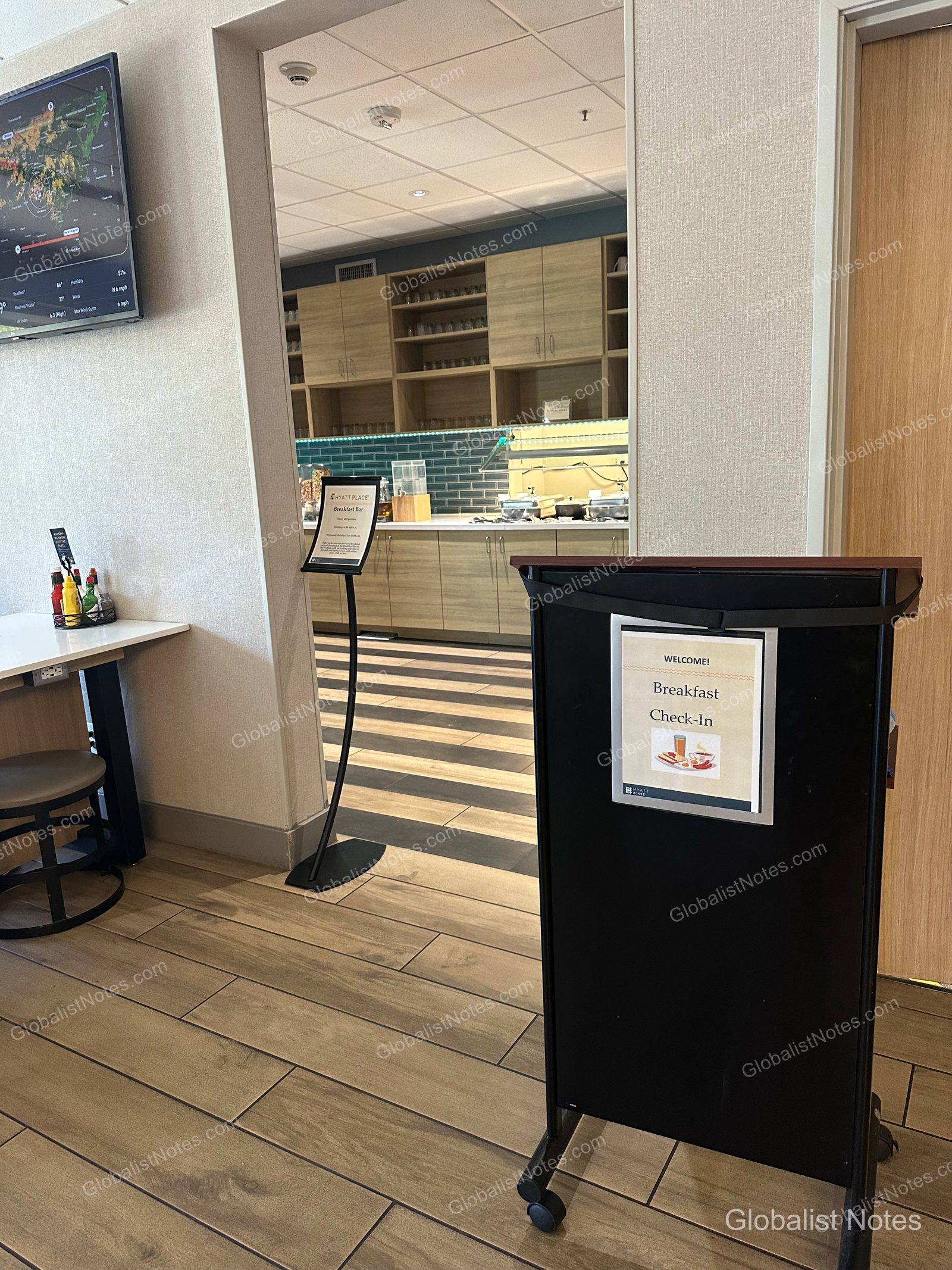 Hyatt Place Flagstaffの朝食エリア入り口。手前に「Breakfast Check-In」のポディウムがあり、奥に朝食カウンターと収納棚が見えます。壁には天気情報が表示されたモニターが設置されています。