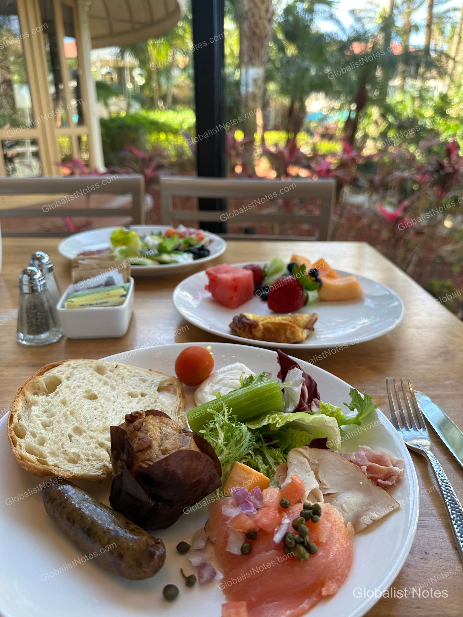 Breakfast table at Hyatt Regency Grand Cypress. The foreground plate features smoked salmon, sausage, a muffin, salad, and bread; the background plate holds fruit and pastries, with a view of the garden through the window.