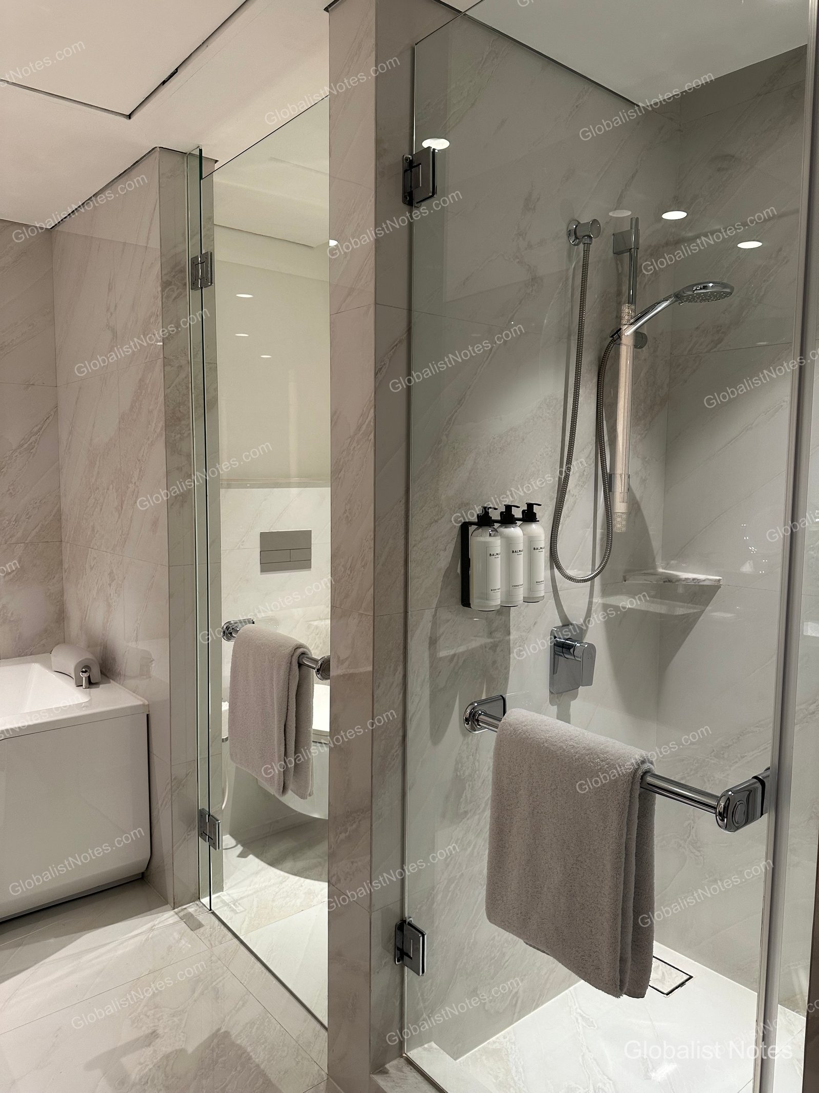 Interior of the Grand Suite bathroom at Grand Hyatt Jakarta, showing marble walls and a glass-enclosed shower and toilet area. The shower features wall-mounted Balmain amenities and neatly hung towels.