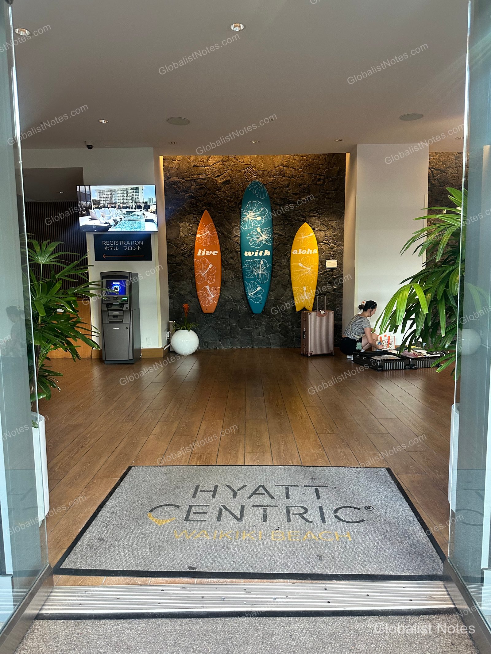 Lobby entrance at Hyatt Centric Waikiki Beach in Honolulu, Hawaii. The floor features a branded logo mat, while the stone wall ahead is decorated with three surfboards inscribed with 'live with aloha.' Check-in monitors and kiosks are positioned to the left.