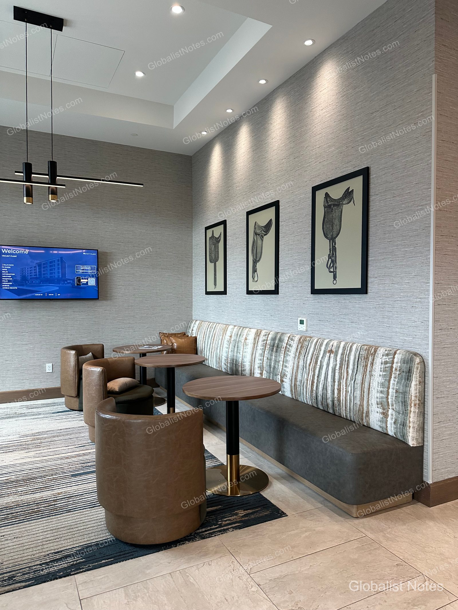 Hyatt Place Bozeman Yellowstone Airportのロビーラウンジ。壁には3つの鞍が描かれたアートが飾られ、ブラウンのレザーチェアやベンチシート、円形テーブルが配されています。左側の壁には案内モニターを設置。