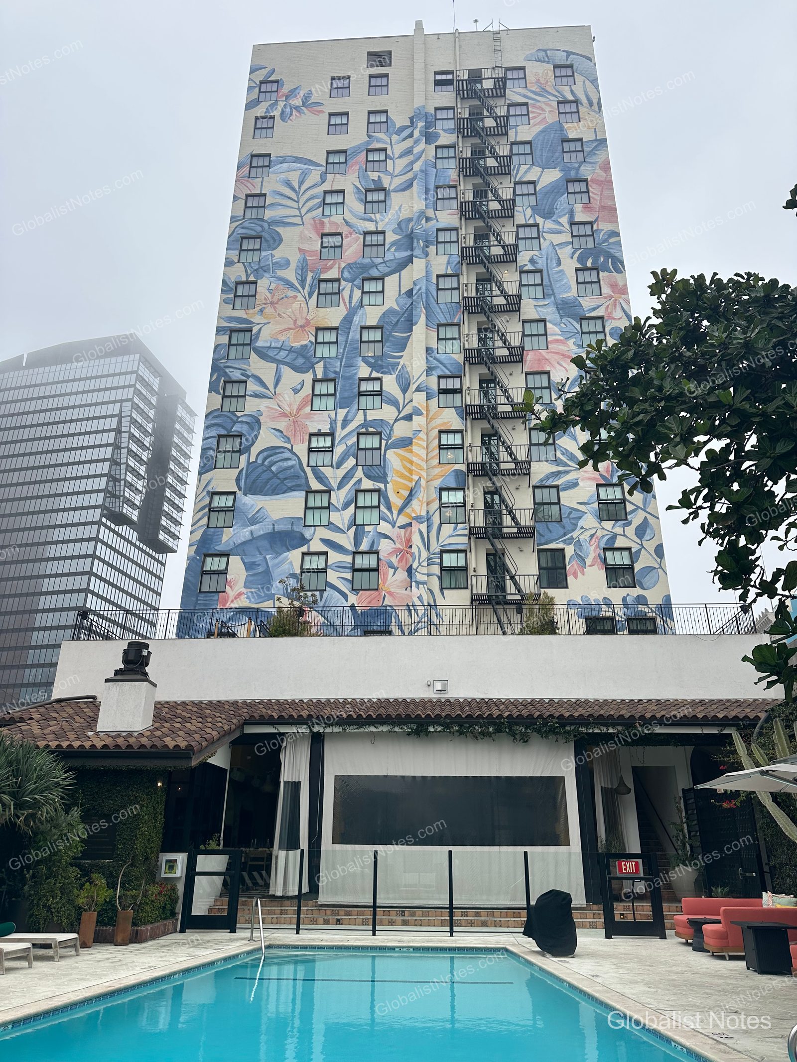 Exterior view of Hotel Figueroa, Unbound Collection by Hyatt in Los Angeles. The building facade features a large mural of blue and pink flowers and leaves, with a rectangular outdoor pool and lounge seating in the foreground.