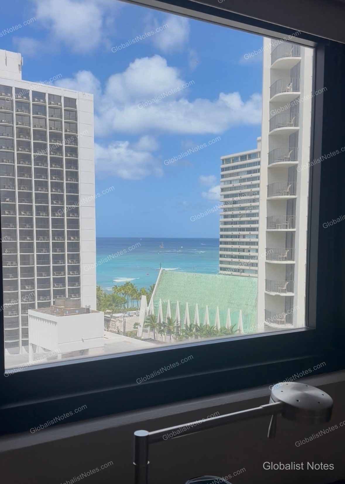 View from the guest room window at Hyatt Place Waikiki Beach, showing tall hotel buildings on either side with a glimpse of the blue ocean, sky, and a building with a green roof in between.