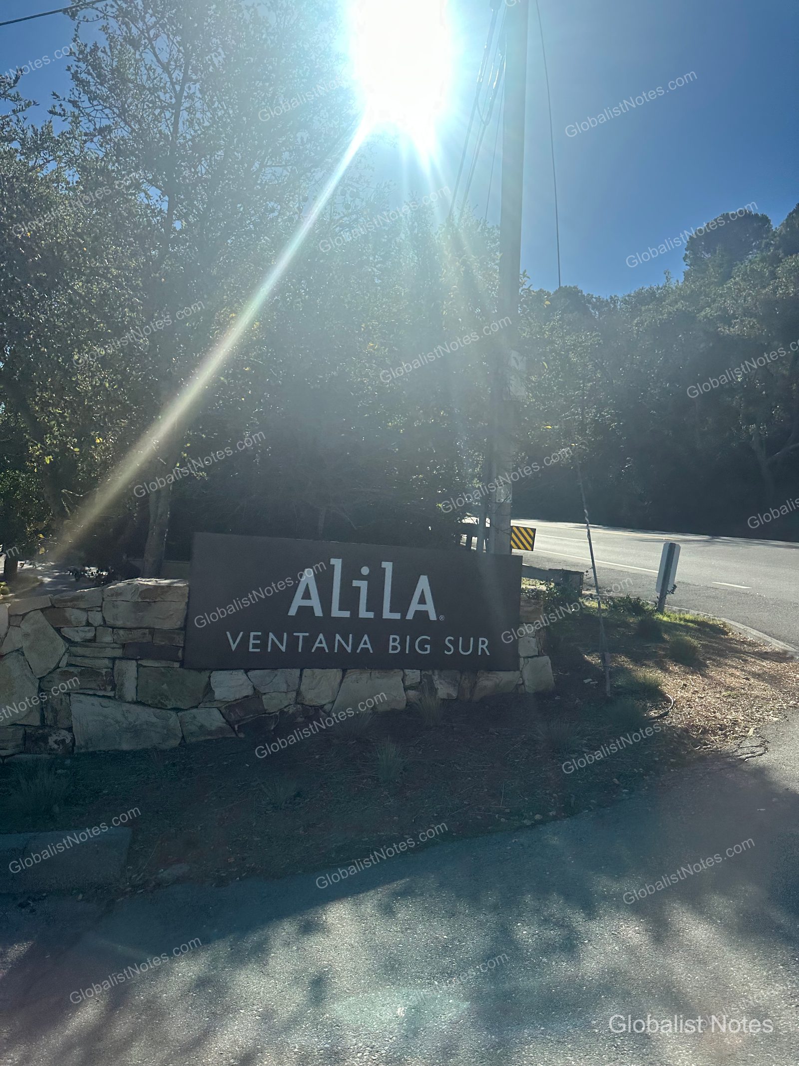 The Alila Ventana Big Sur entrance sign mounted on a stone wall, with dense trees and a road in the background under bright sunlight.