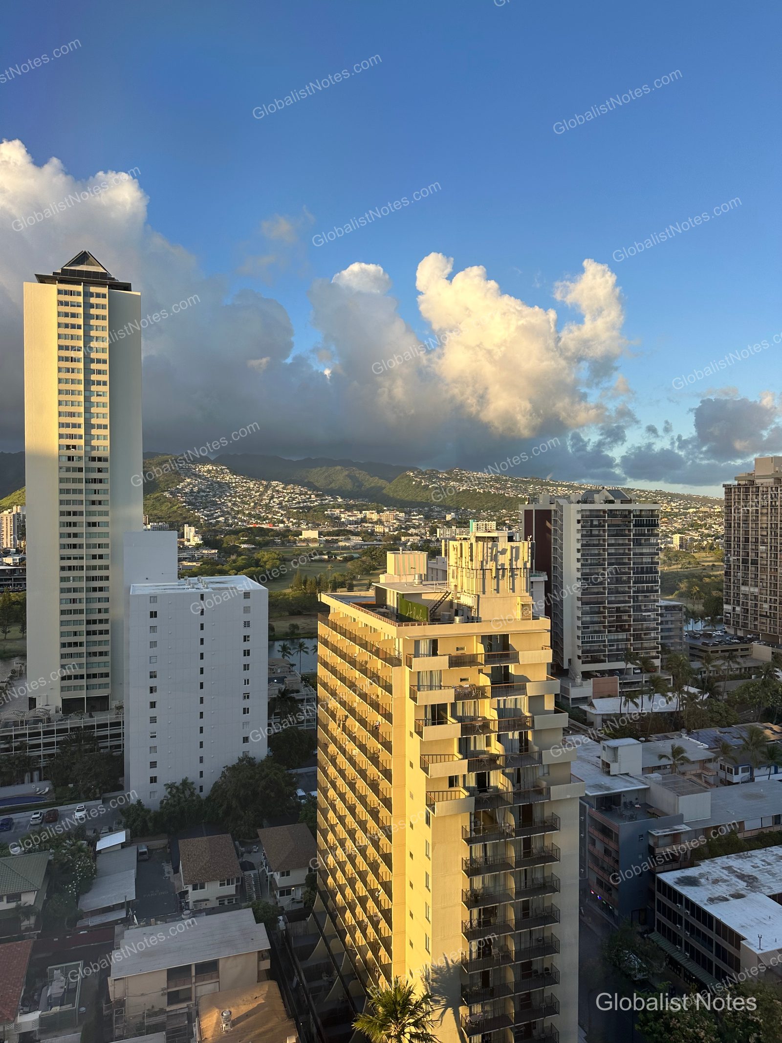 View of the Honolulu cityscape from a guest room at Hyatt Centric Waikiki Beach, featuring dense high-rise buildings set against a backdrop of green mountains and a cloudy sky under afternoon light.