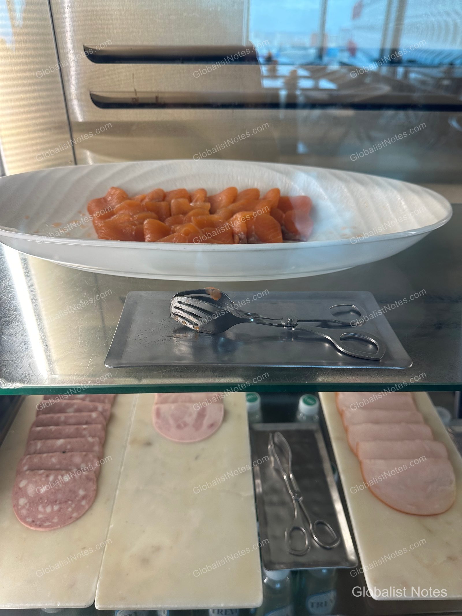 Inside the refrigerated showcase at the Grand Hyatt Jeju breakfast buffet. The top white oval platter holds smoked salmon, while the bottom marble shelf features two types of sliced ham, each with serving tongs provided.