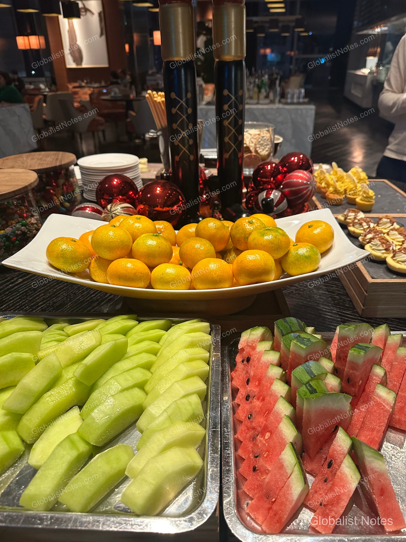 A buffet table at the Grand Hyatt Jeju lounge featuring plates of fresh fruit including tangerines, melon, and watermelon, accented with red Christmas ornaments and tall cylindrical decor.