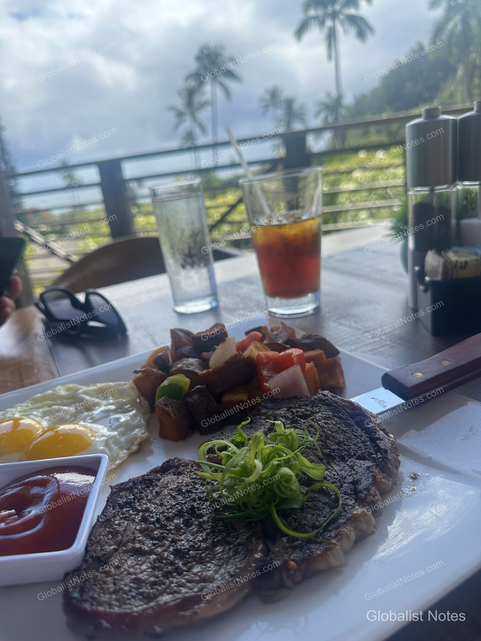 A breakfast plate with steak, fried eggs, and roasted potatoes on an outdoor terrace table at Hana-Maui Resort, with iced drinks and a backdrop of Maui's palm trees and natural scenery.