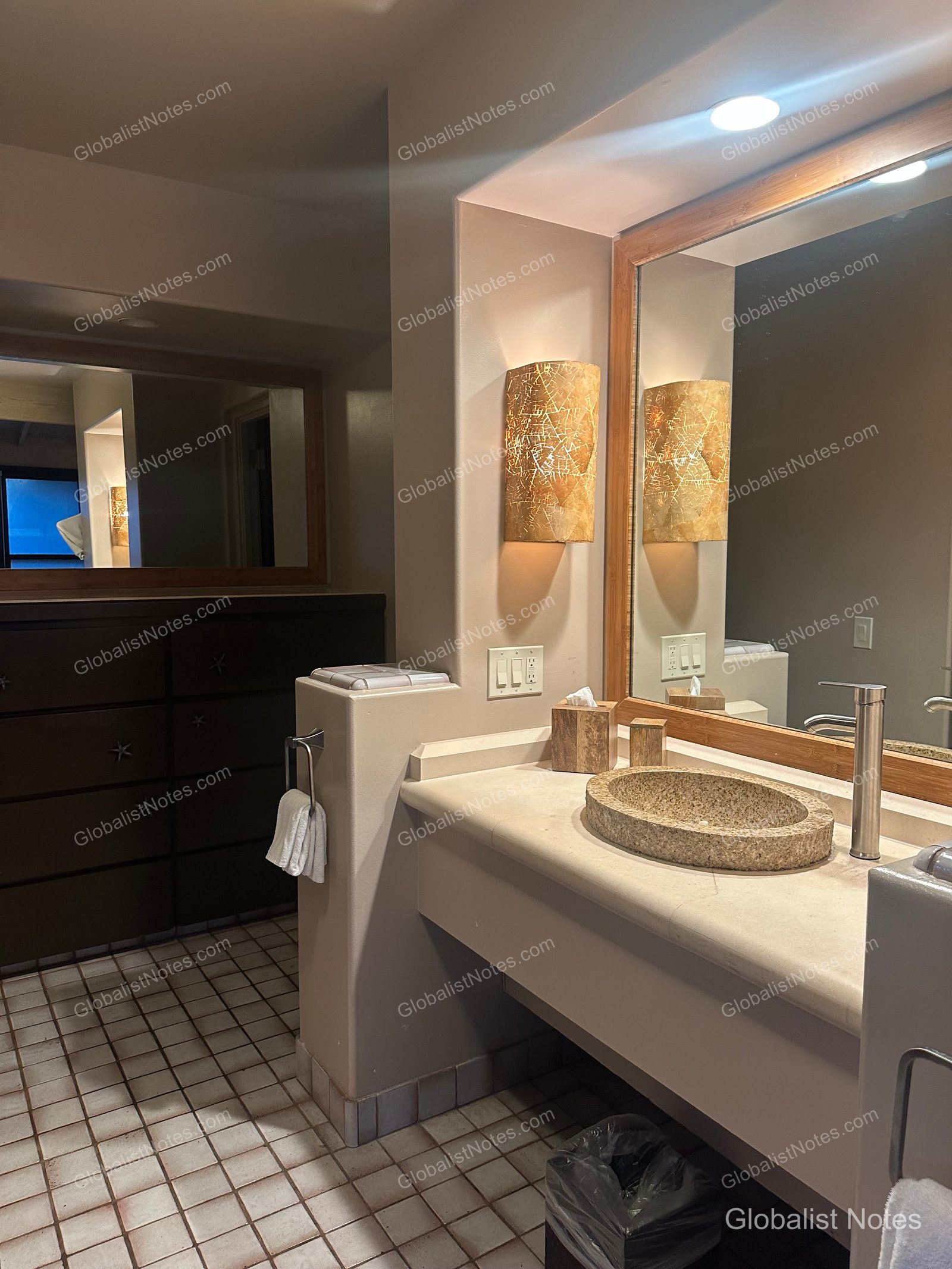 Bathroom interior of a 1 King Bed Ocean View Bungalow at Hana-Maui Resort. It features a stone-textured circular sink, wall sconces providing warm lighting on both sides of the mirror, tiled flooring, and dark-toned cabinetry along the wall.