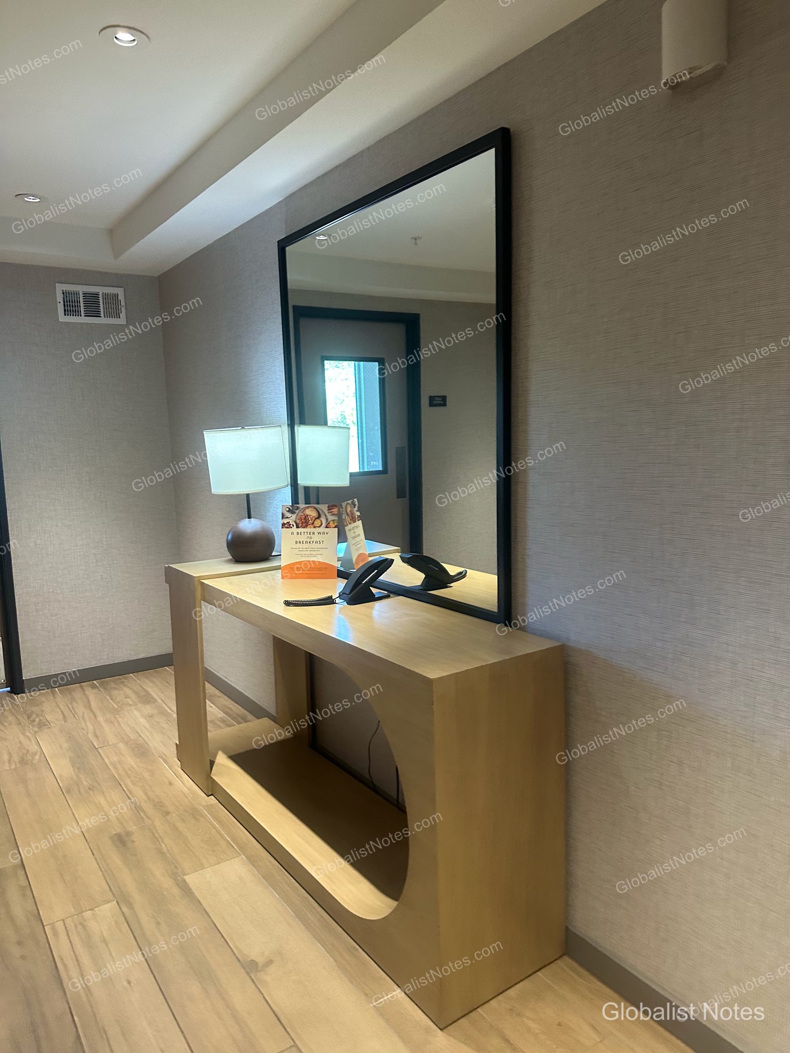 Hyatt Place Flagstaffの客室にある明るい木目調のコンソールテーブル。照明、電話、朝食の案内が置かれ、壁には大きな黒いフレームの鏡が掛けられています。床は木目調の仕上げです。