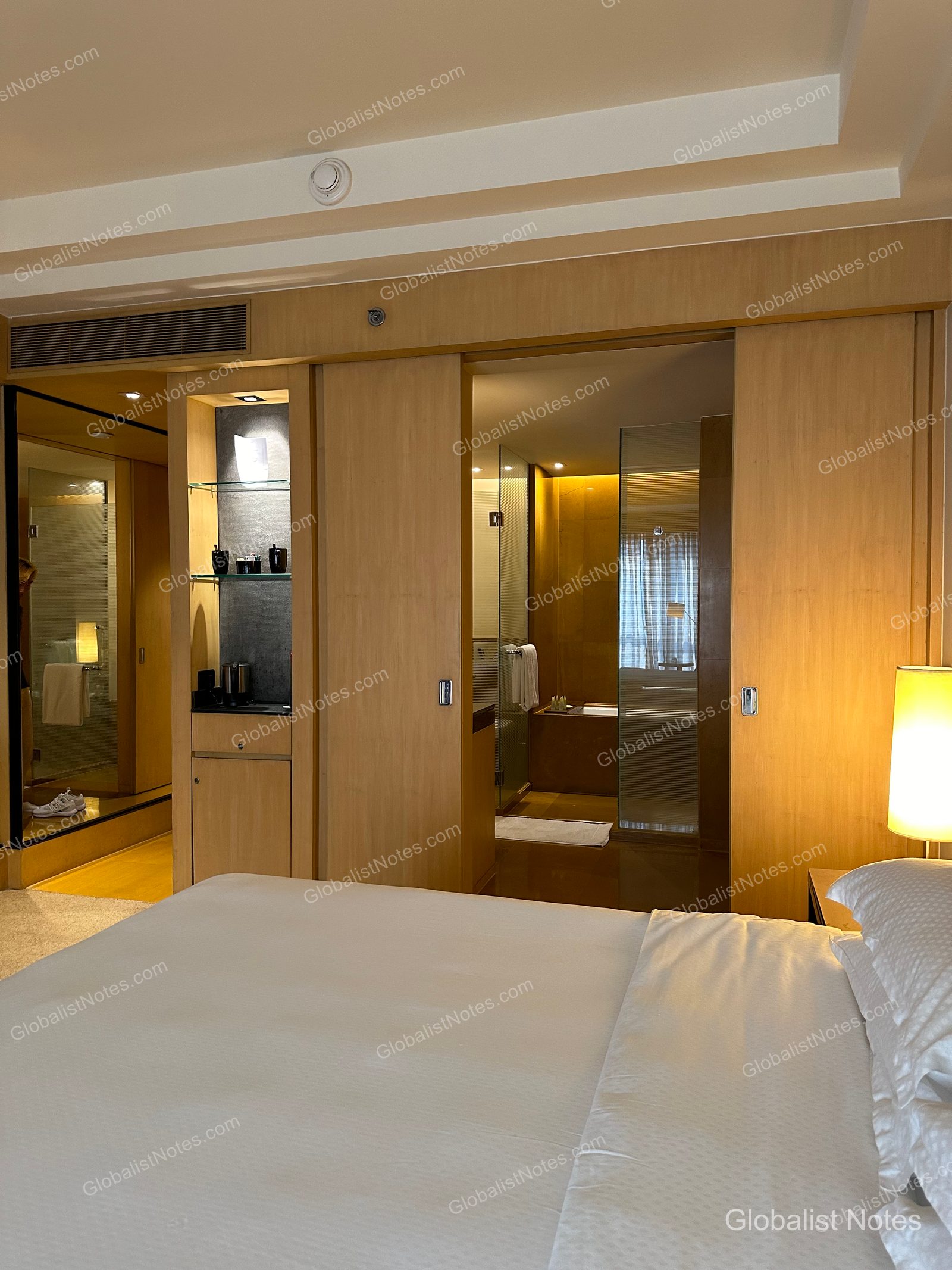 Interior view of a room at Grand Hyatt Mumbai Hotel and Residences, showing a sliding door open to the bathroom and minibar area, with light-toned wood paneling, illuminated shelving, and a bathtub in the background.