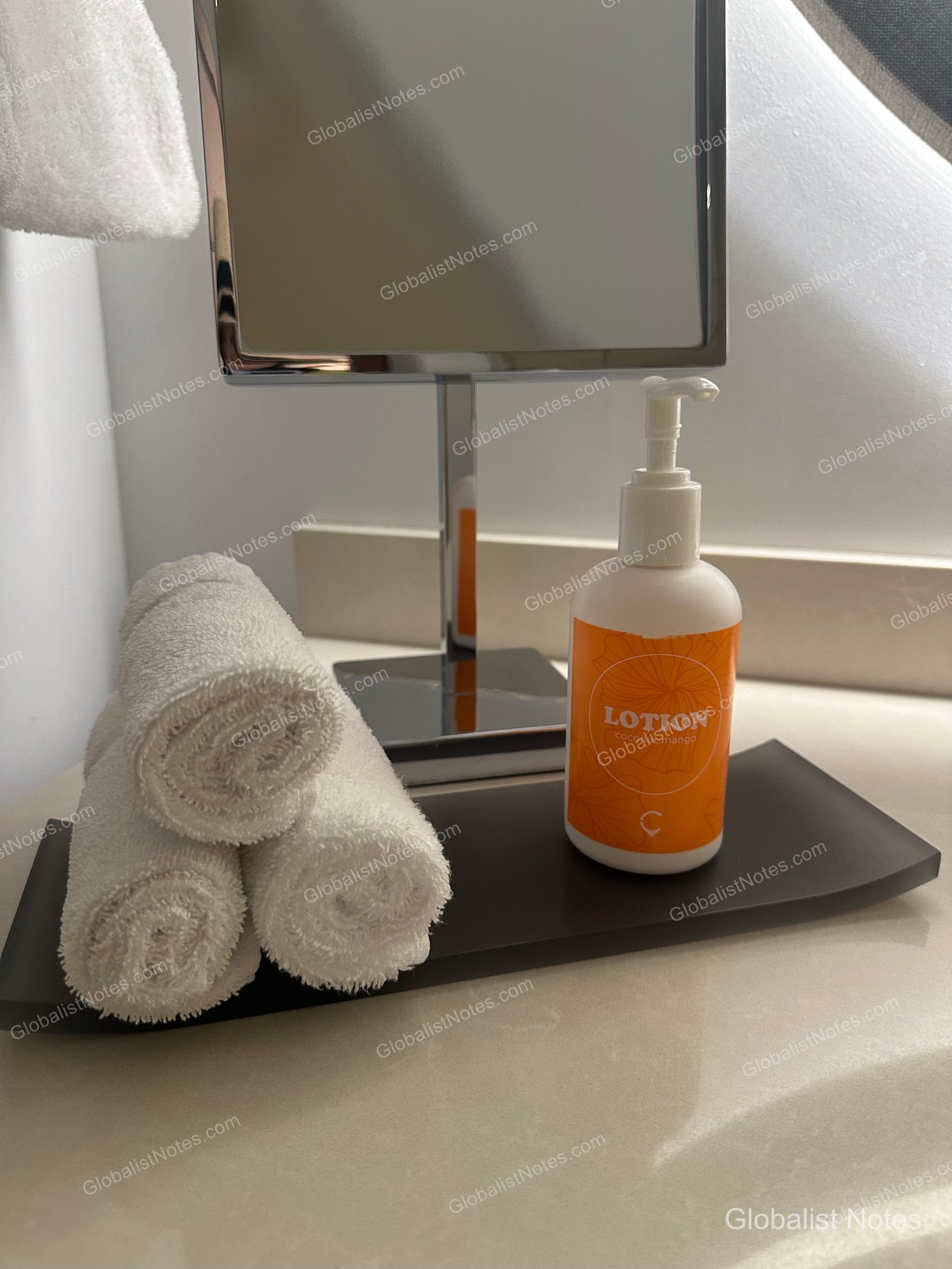 A dark brown tray on the bathroom vanity at Hyatt Centric Waikiki Beach holds three neatly rolled white hand towels and a bottle of Coco Mango body lotion with an orange label, with a square tabletop mirror positioned behind them.