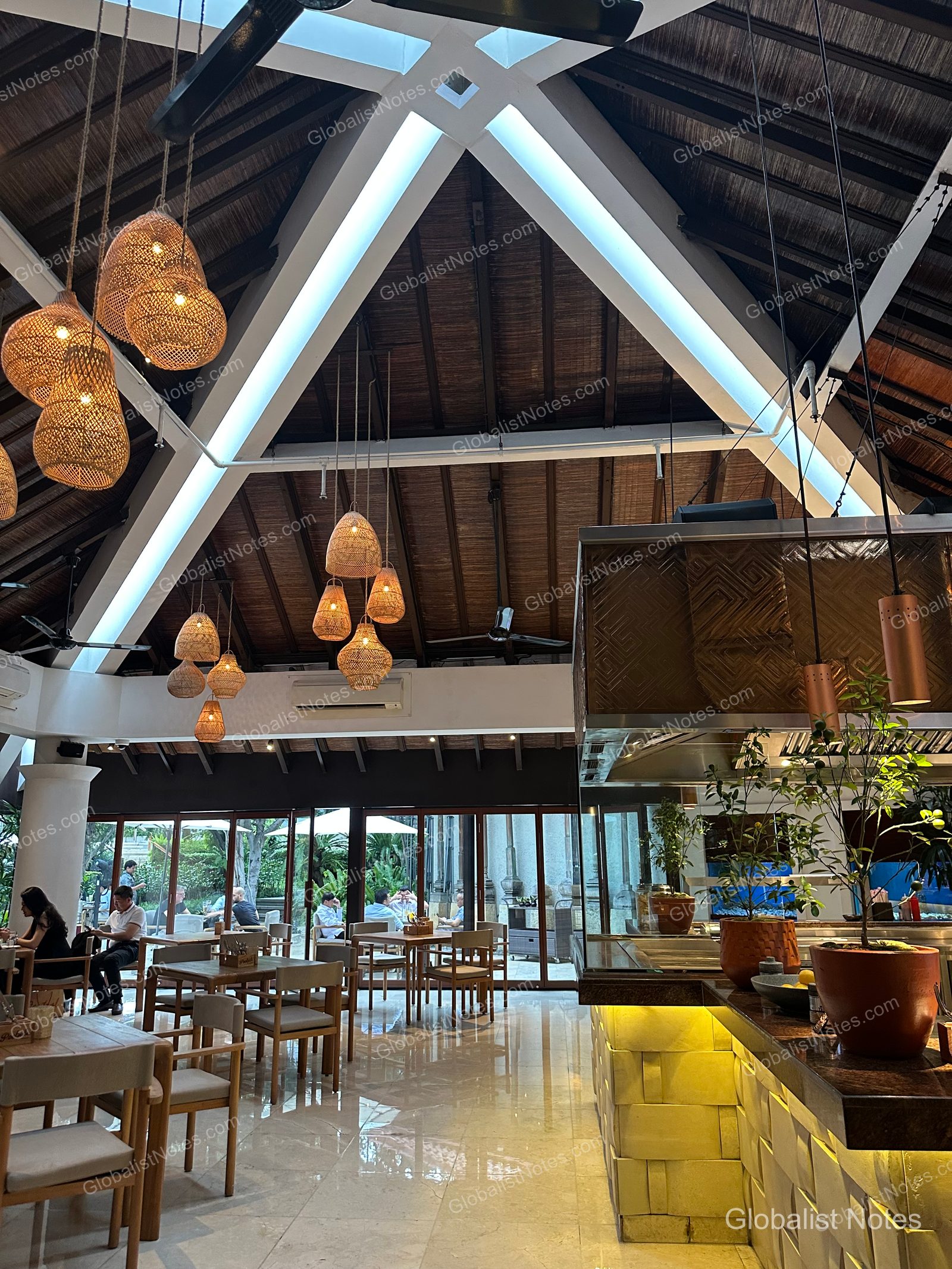 Interior dining space at Grand Hyatt Jakarta with rattan pendant lights hanging from a high gabled roof, wooden tables and chairs, a service counter, and floor-to-ceiling windows looking out onto the garden and guests dining outside.