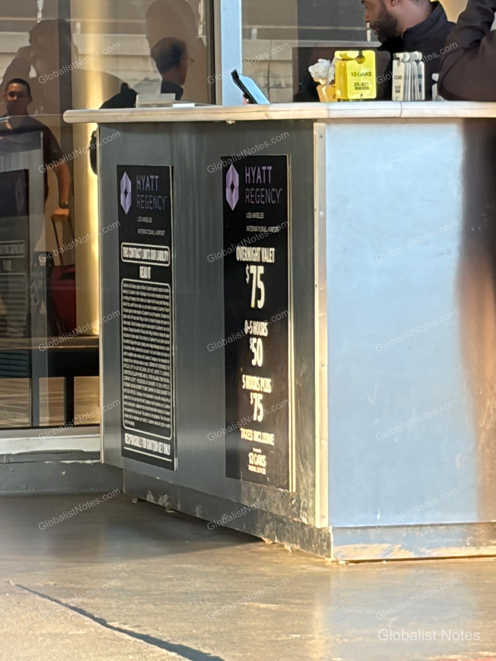Outdoor valet parking desk at Hyatt Regency Los Angeles International Airport. The black signage displays detailed hourly parking rates, including the $75 overnight valet fee, with a staff member standing behind the desk.