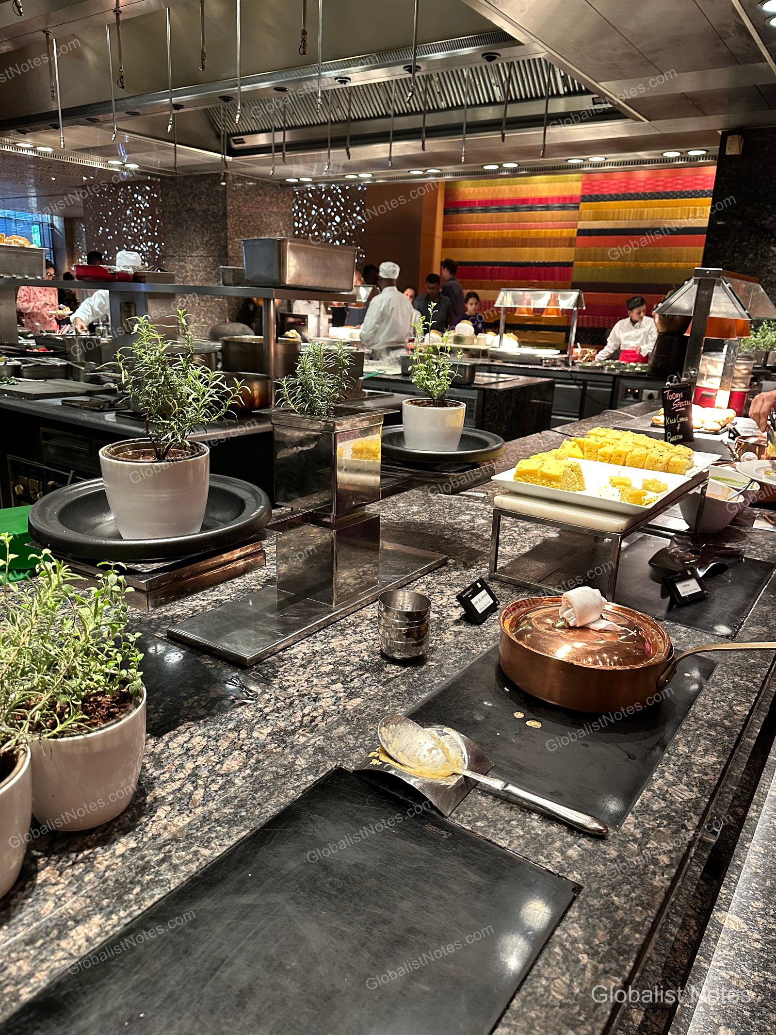 A live cooking station at the Grand Hyatt Mumbai breakfast buffet, with marble countertops displaying herb pots, plates of dhokla, and copper pots, as chefs work in the background.
