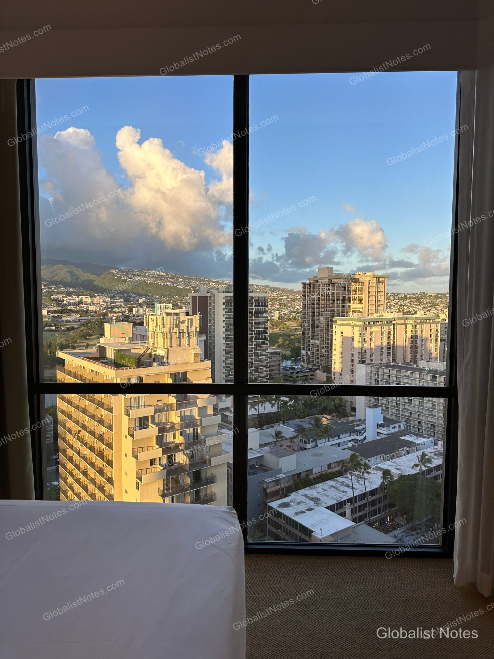 Afternoon sunlight hits the Honolulu city buildings and distant mountain range, viewed through the floor-to-ceiling windows of a guest room at Hyatt Centric Waikiki Beach. A portion of a white bed frame is visible in the foreground under a partly cloudy blue sky.