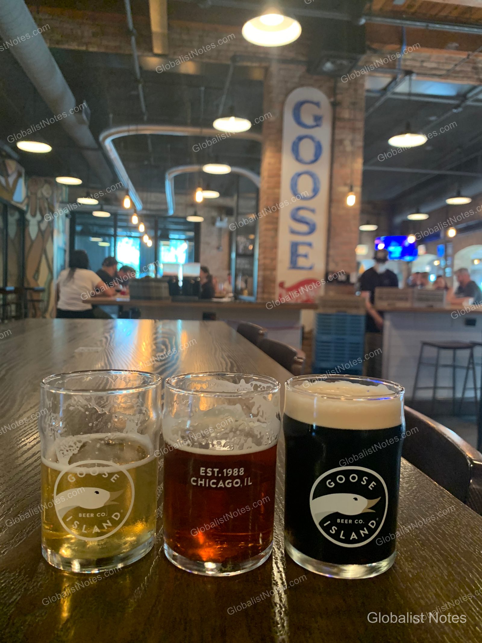 Goose Island Breweryのテーブルに並ぶ3種類のビールサンプラー。背景には高い天井やレンガの柱、賑わう店内の様子が写っている。
