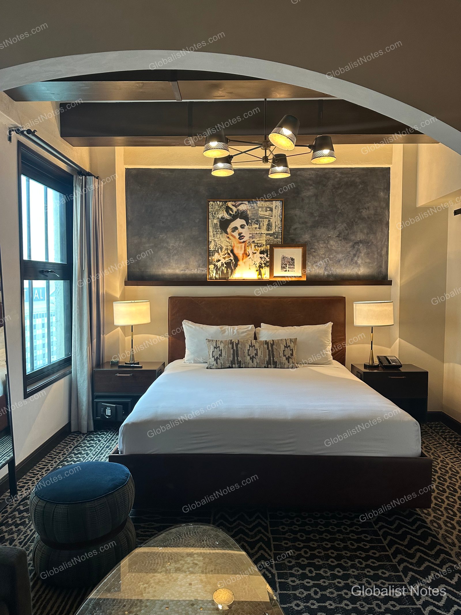 Interior of a guest room at Hotel Figueroa in Los Angeles, featuring a queen-size bed under an arched ceiling. A portrait hangs above the headboard, a modern chandelier is suspended from the ceiling, and a partial view of the city is visible through the window on the left.