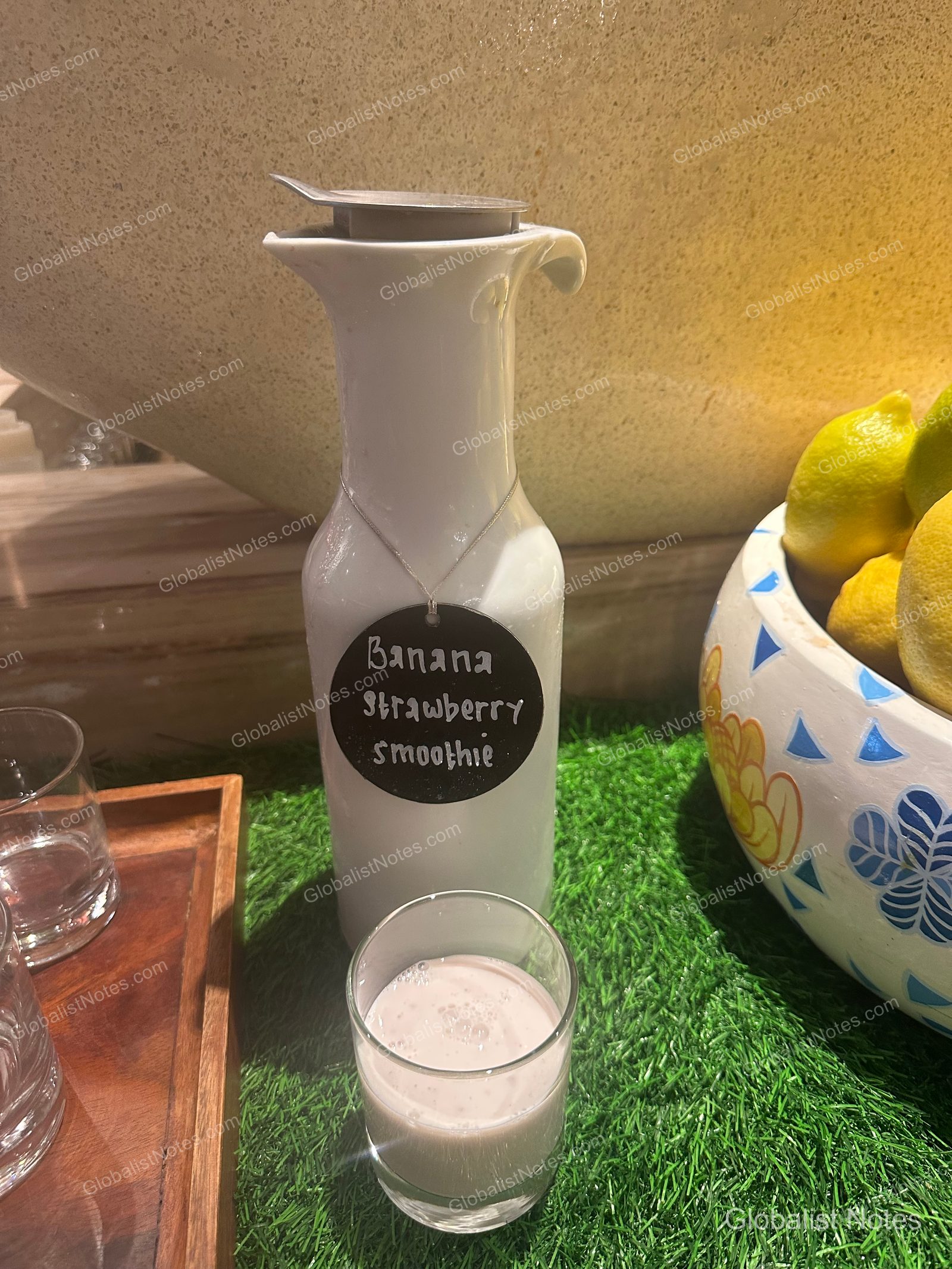 A white bottle of banana strawberry smoothie, a glass, and a bowl of lemons displayed on artificial turf at the Grand Hyatt Jakarta breakfast buffet.