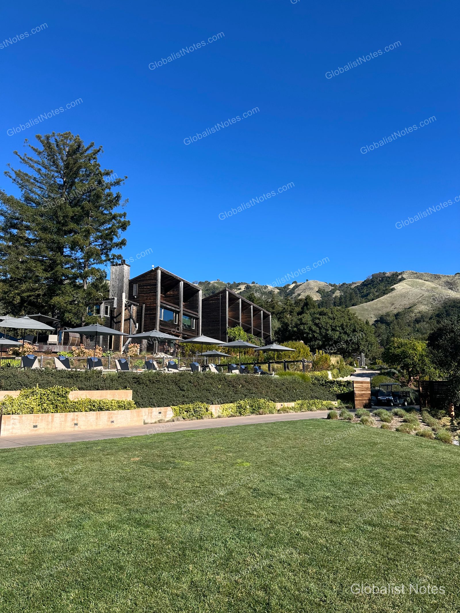 A panoramic view of the Alila Ventana Big Sur resort, showing the wooden guest buildings, outdoor pool, and umbrellas across a green lawn under a clear sky.