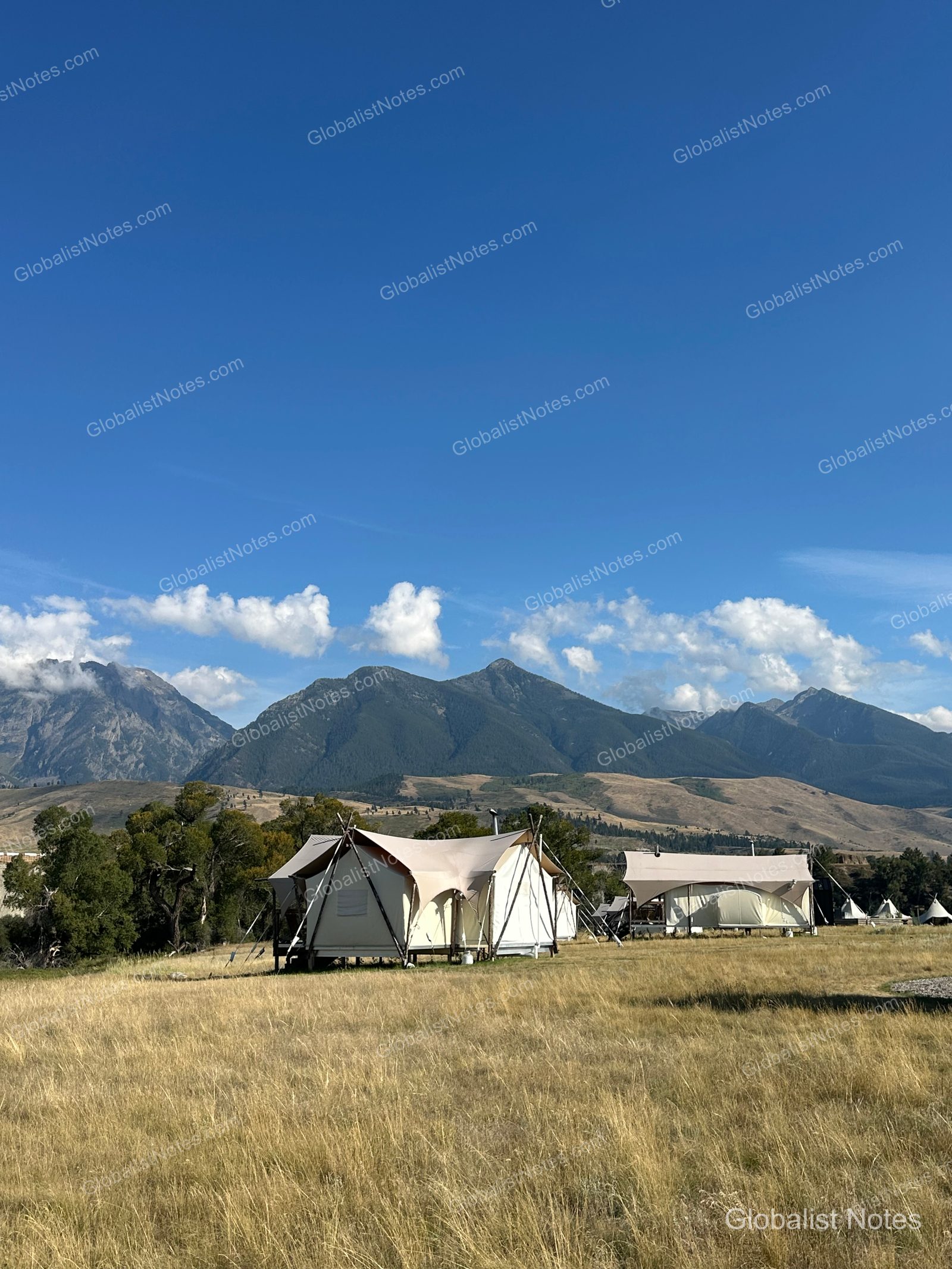 Under Canvas North Yellowstone - Paradise Valleyの全景。黄金色の草原に佇むテントの背後には、澄んだ空と雄大な山脈が広がっています。