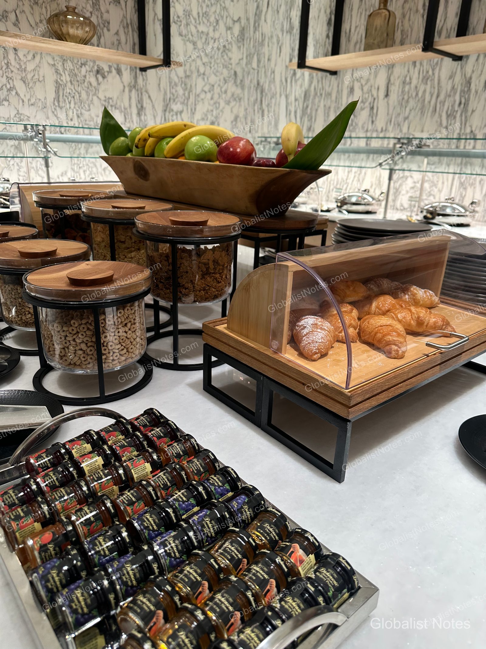 Hyatt Regency Newport Beach 早餐自助餐一景。大理石桌面上整齊陳列著玻璃罐裝麥片、木碗盛裝的蘋果與香蕉，以及蓋有透明罩的羊角麵包與各式小瓶果醬。