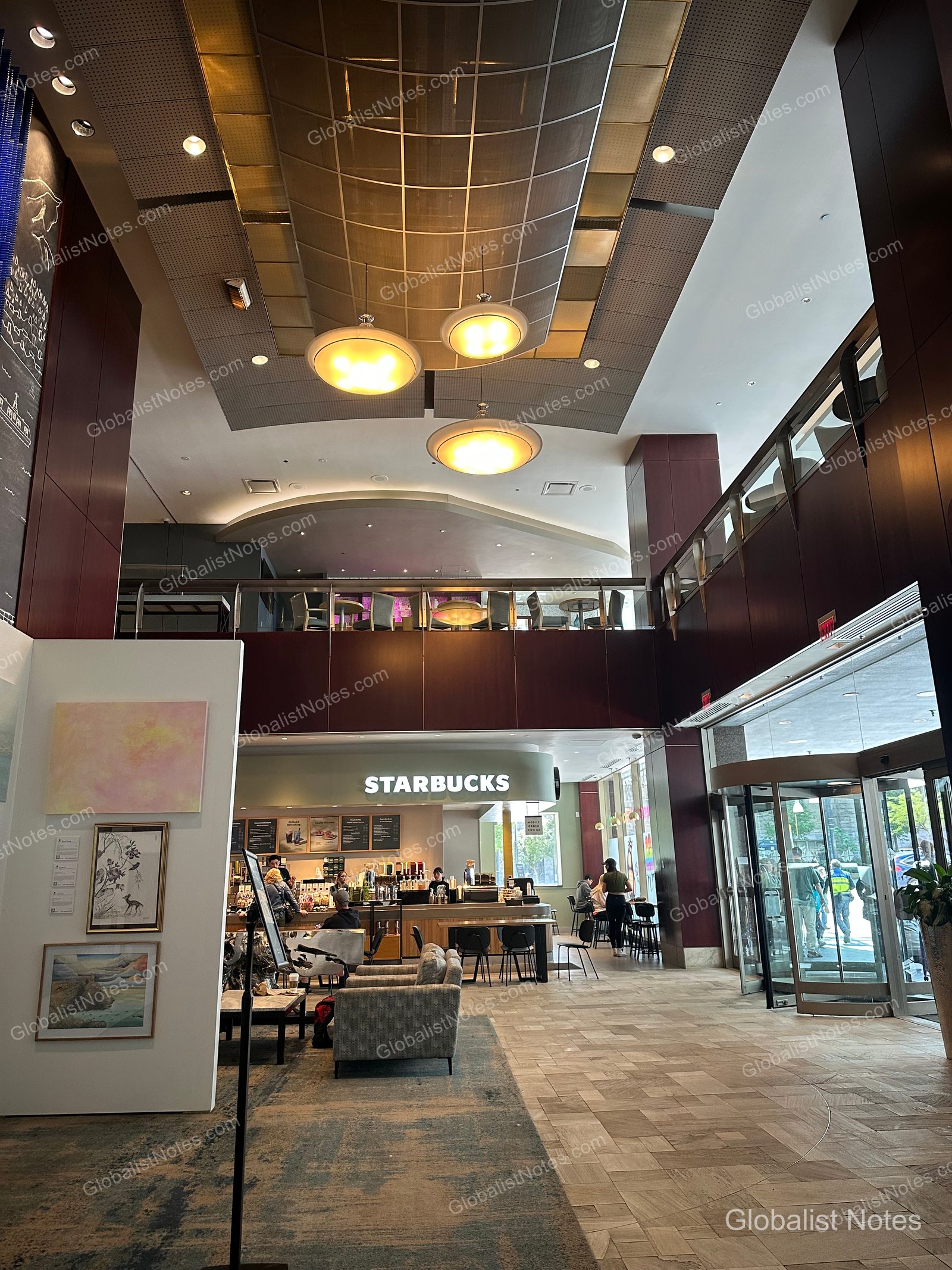 View of the high-ceilinged lobby at Hyatt Regency Vancouver. The center features warm lighting and seating, with a Starbucks located on one side and revolving doors leading to the exterior on the right.