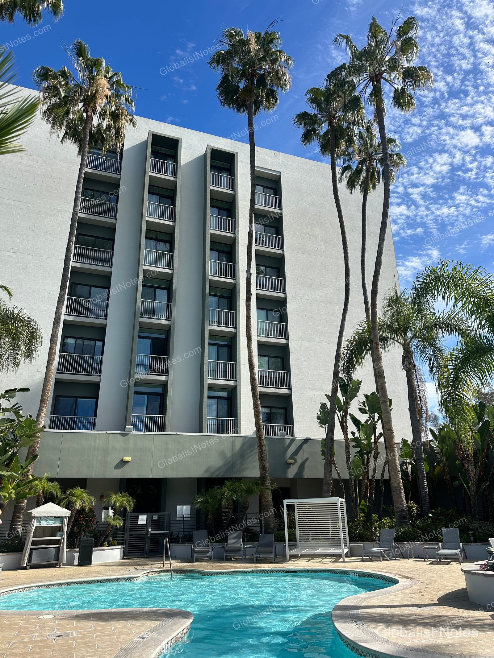 位於 Newport Beach 的 Hyatt Regency Newport Beach West 戶外泳池，背景為飯店建築。藍天映襯下種植著一排棕櫚樹，泳池周邊配置躺椅與涼亭。