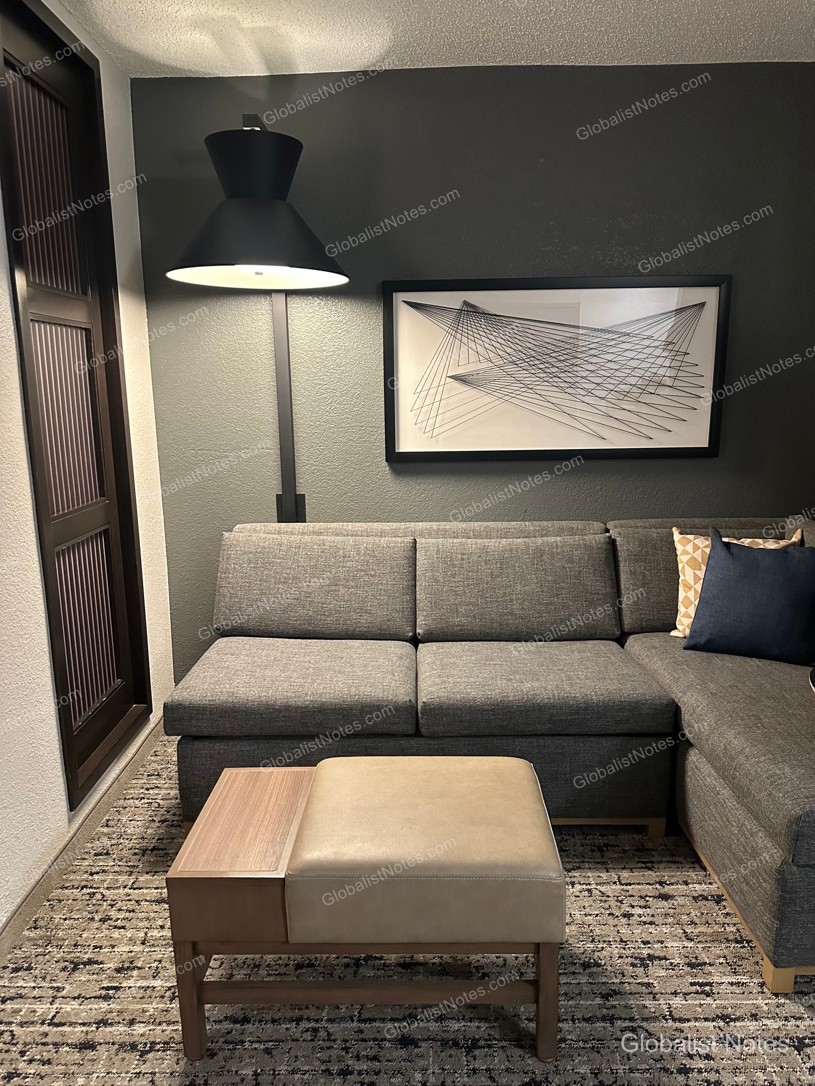 Hyatt Place El Paso Airport Junior Suite 的起居空間。深灰色牆面前擺放灰色轉角沙發與米色腳凳，牆上掛有幾何線條畫作，現代風格的黑色燈具照亮沙發區。