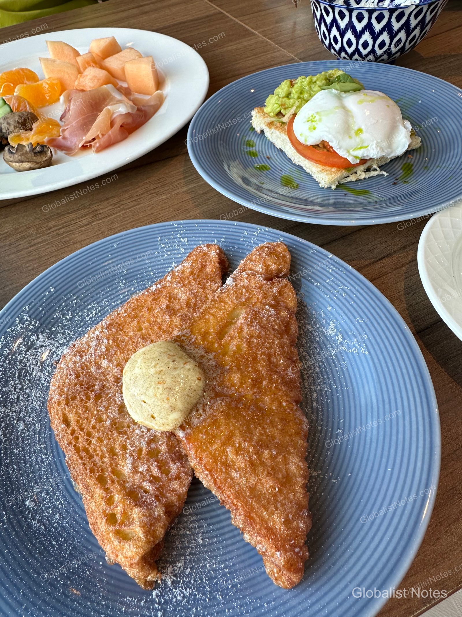 A breakfast spread at Hyatt Regency Malta featuring a plate of melon, tangerines, and jamón, alongside avocado toast with a poached egg and powdered sugar-dusted French toast.
