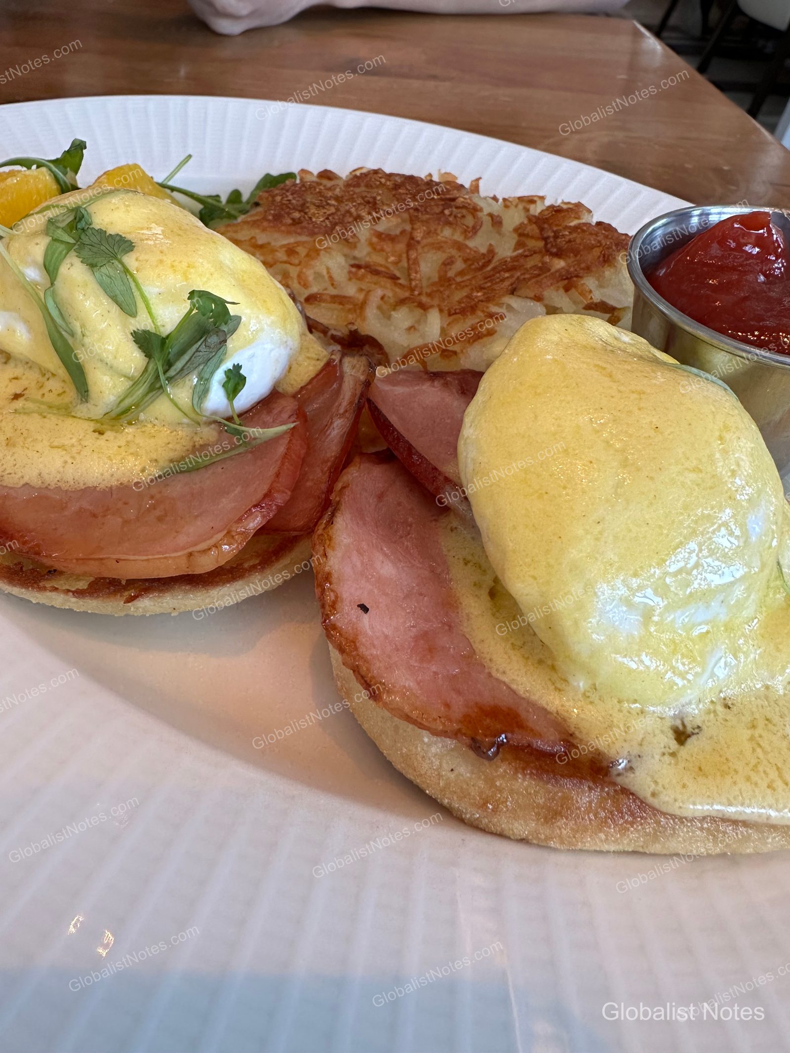 Two eggs Benedict on a white plate with hash browns and a small metal container of ketchup. The dish features English muffins topped with ham, poached eggs, and hollandaise sauce, garnished with green herbs.