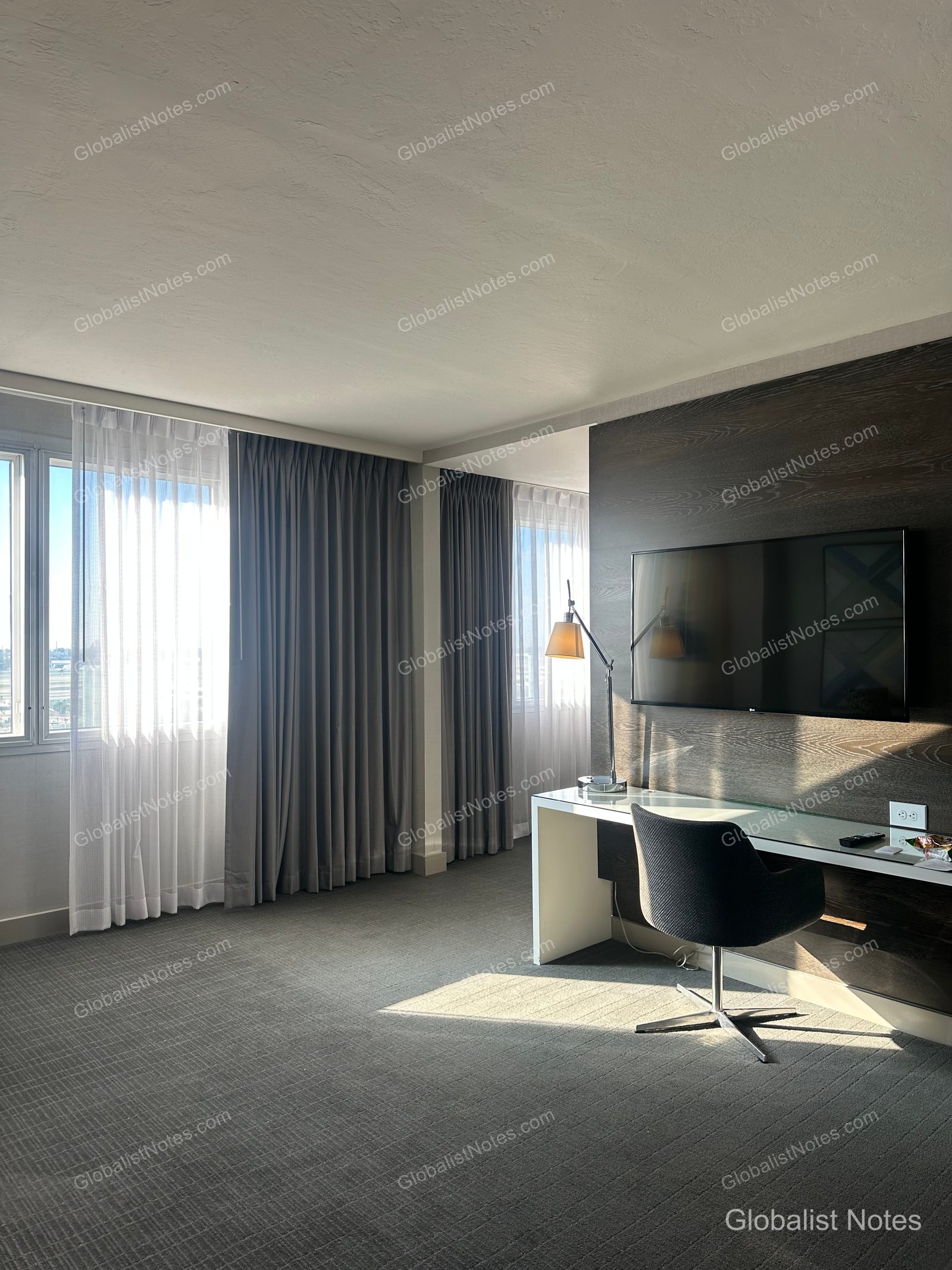 Interior of a One Bedroom King Suite at Hyatt Regency Los Angeles International Airport. The room features a large TV and a work desk against the wall, with carpeted flooring throughout. Gray curtains hang by the windows, allowing natural light into the room.