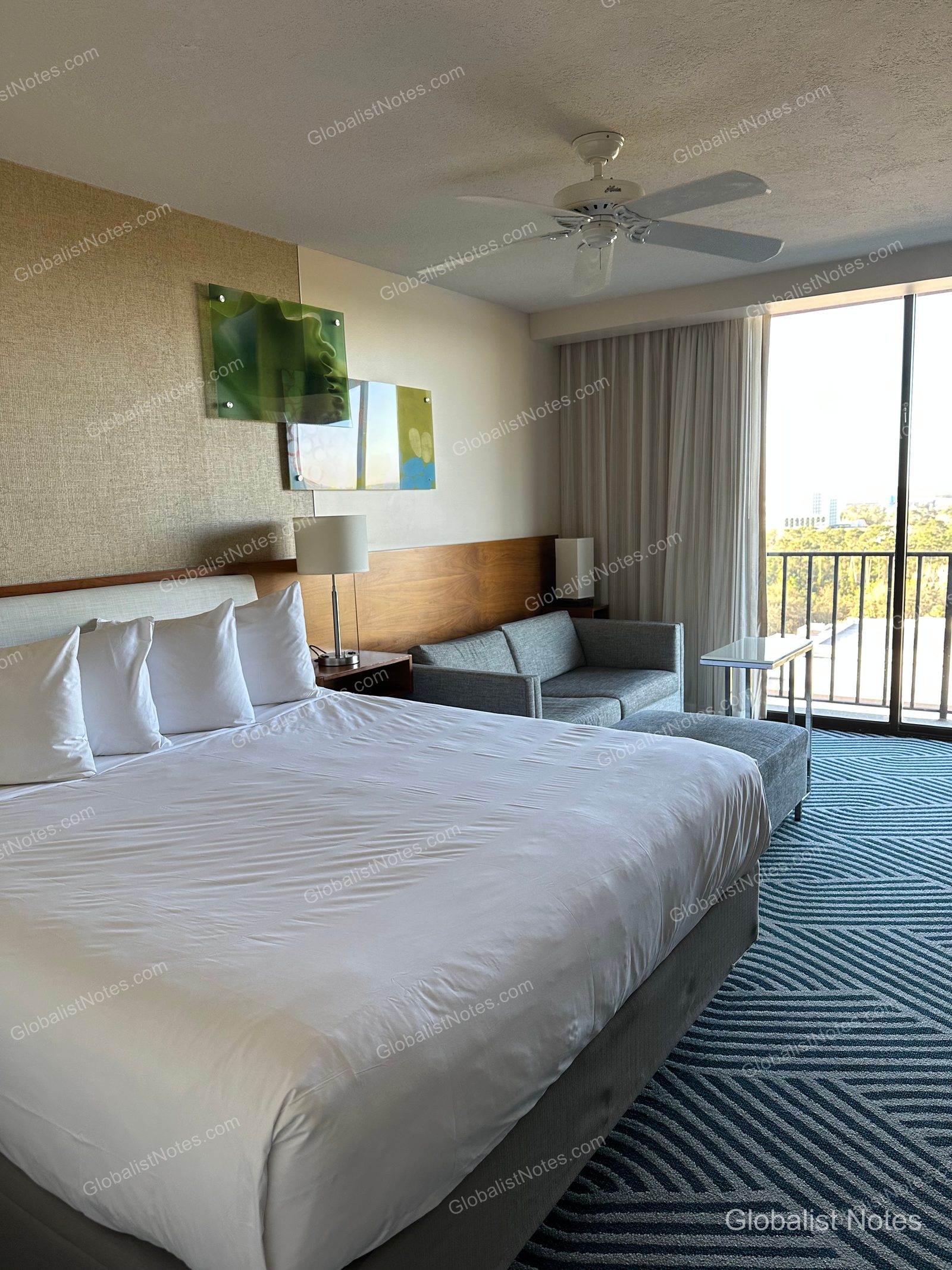 Interior of a standard king room at Hyatt Regency Grand Cypress. The room features a king bed with white linens, a gray sofa to the side, floor-to-ceiling windows leading to a balcony, and geometric patterned carpeting.