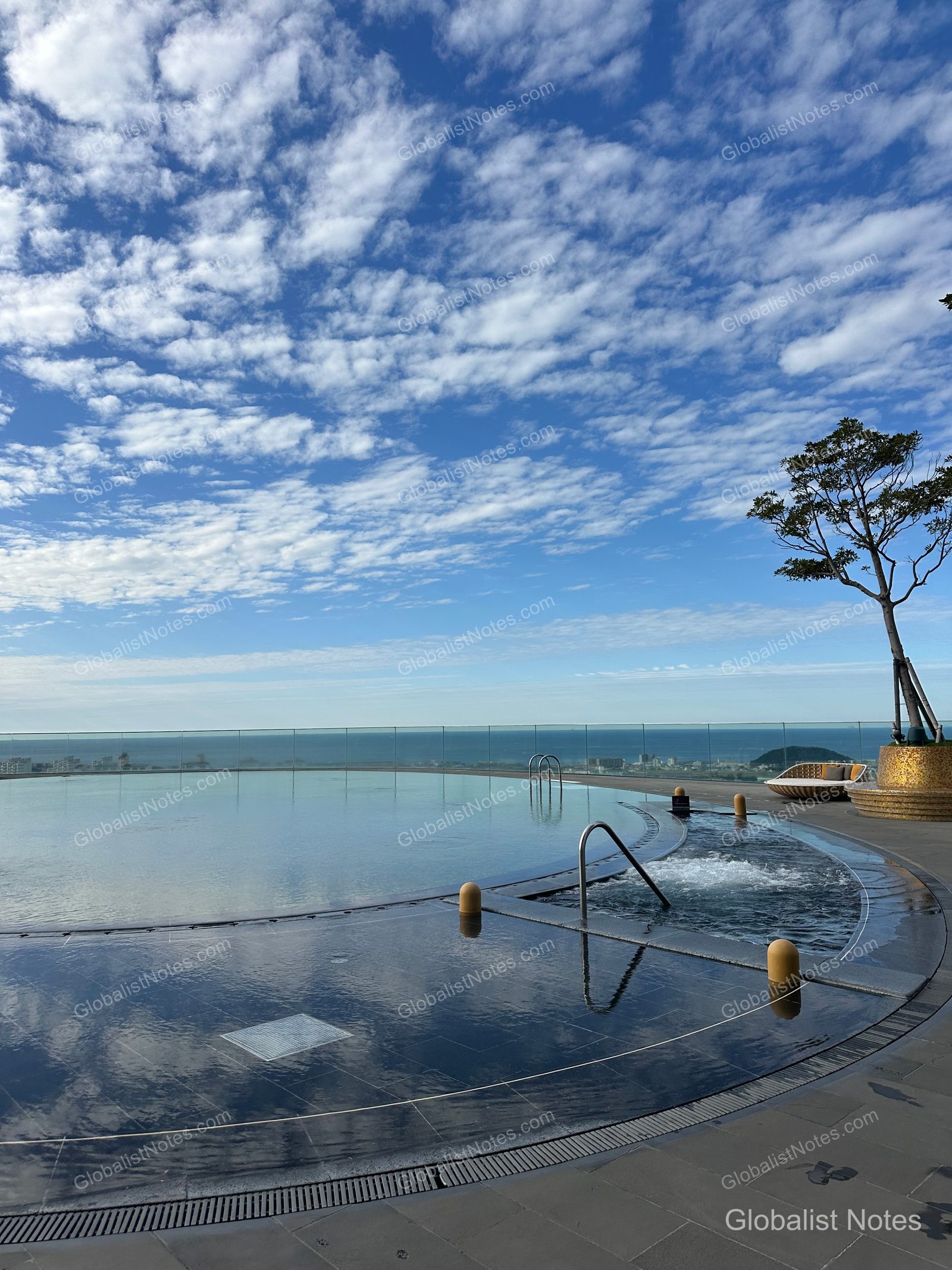 View of the outdoor infinity pool at Grand Hyatt Jeju, with the clear autumn sky and clouds reflected on the water and the Jeju sea stretching beyond the horizon. A small tree and lounge area are located next to the pool.